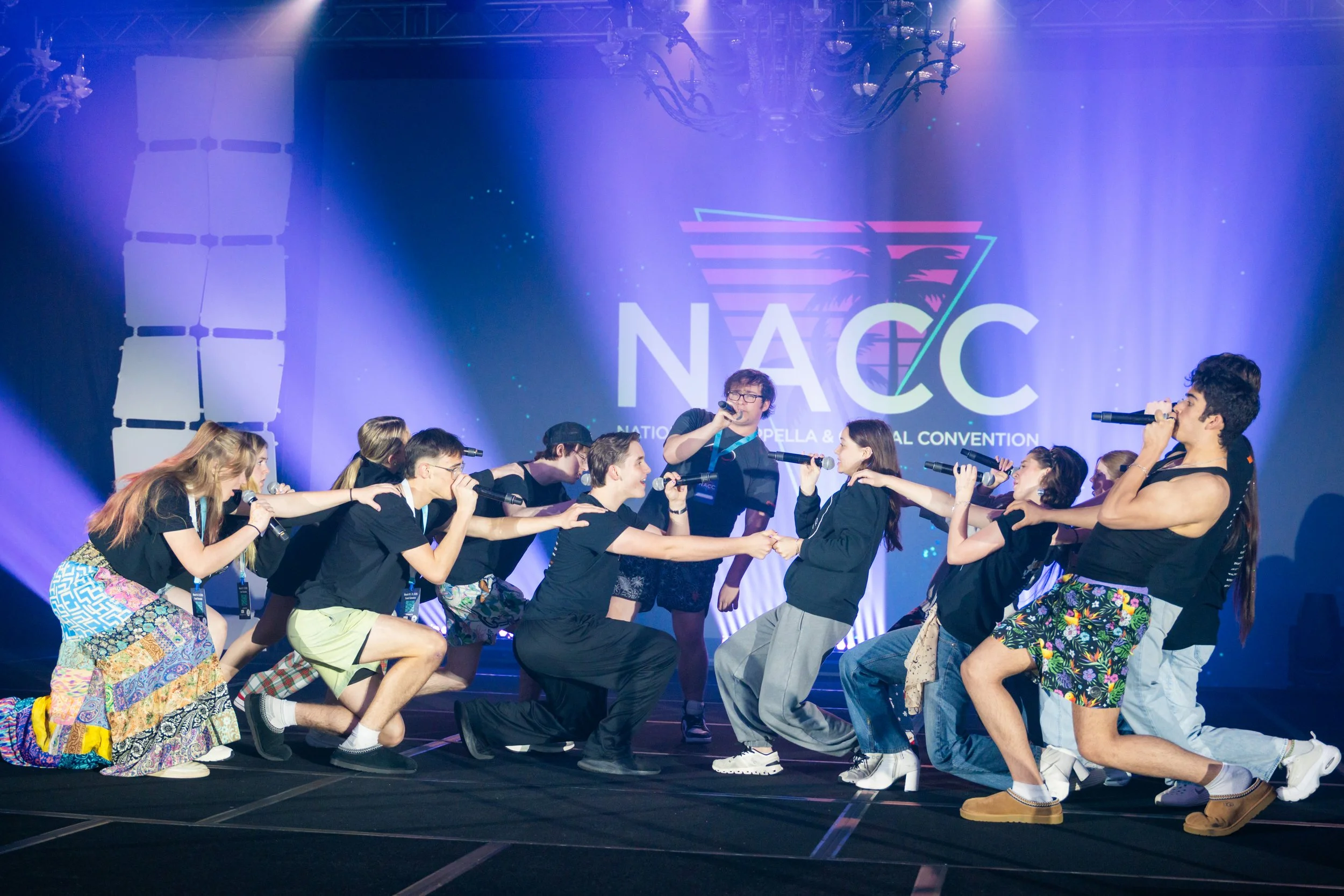 A group of young people participating in a choreographed activity on stage at the NAC (National Anime Convention), with a large logo and name projected on the background screen. Some are kneeling, holding hands, while others are standing and singing 