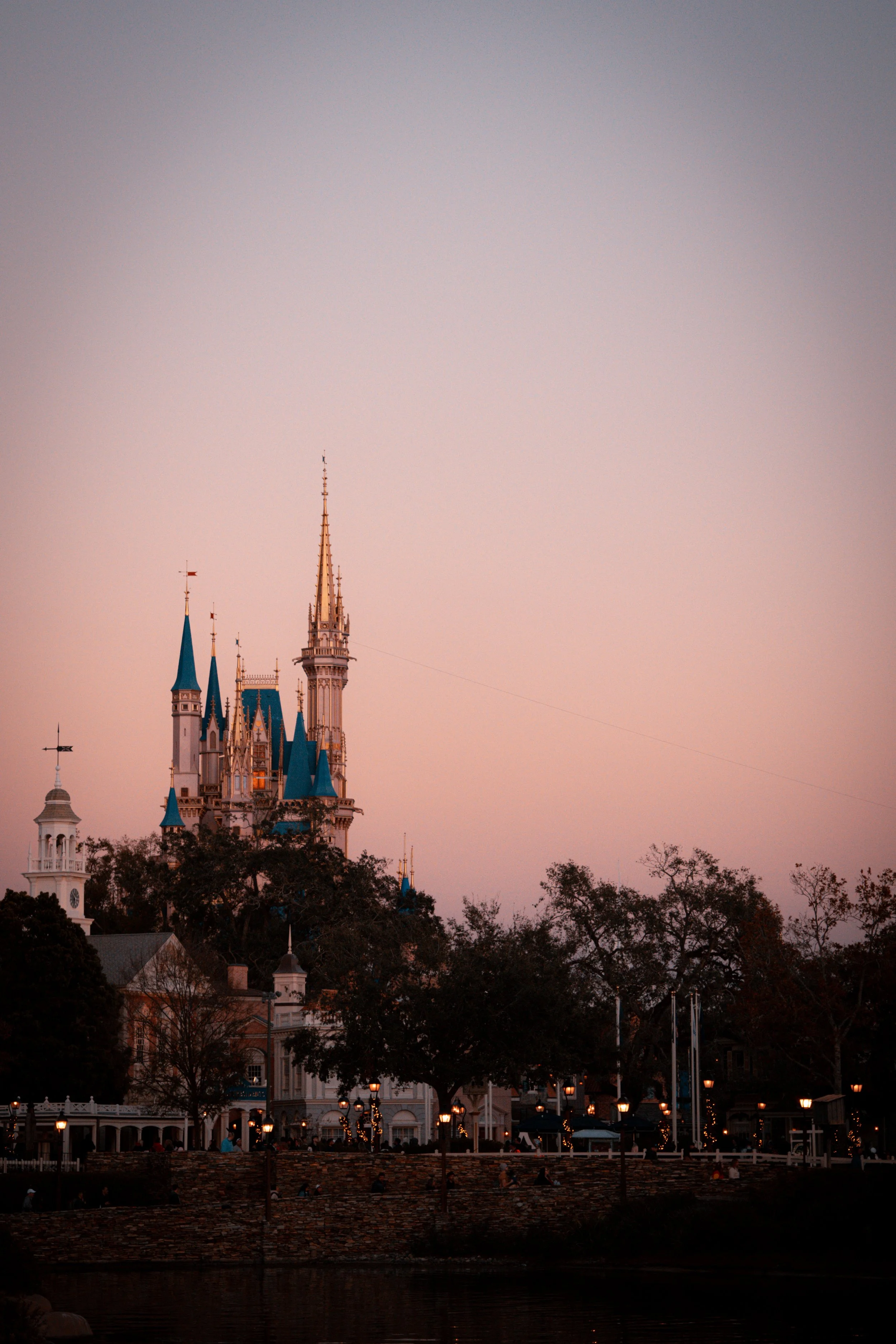 Disney castle at sunset with pink and purple sky, surrounded by trees and lit lanterns.
