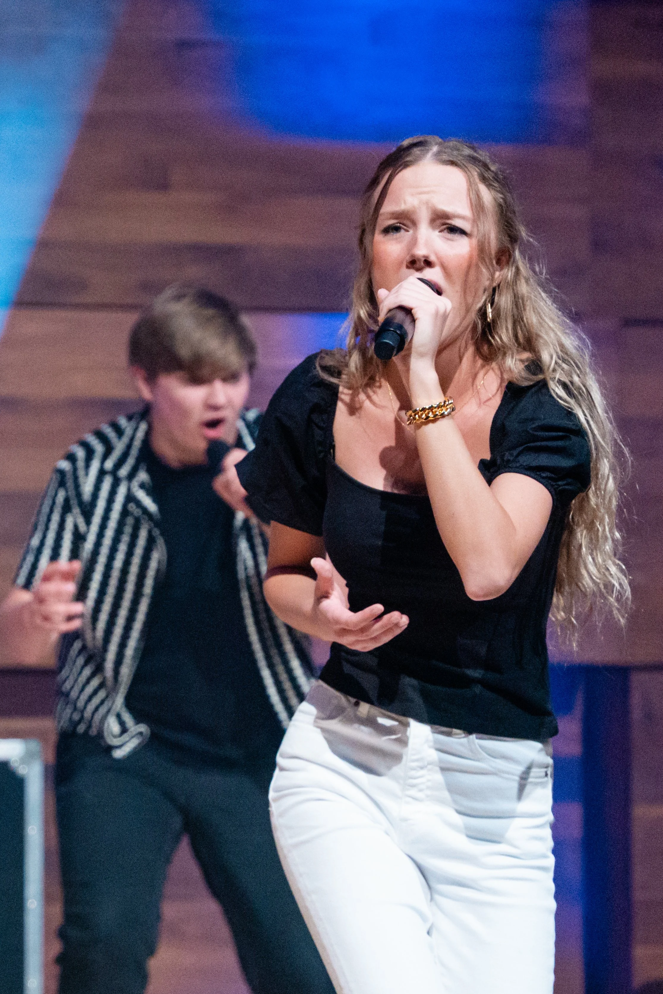 A woman singing into a microphone on stage with a young man in the background.