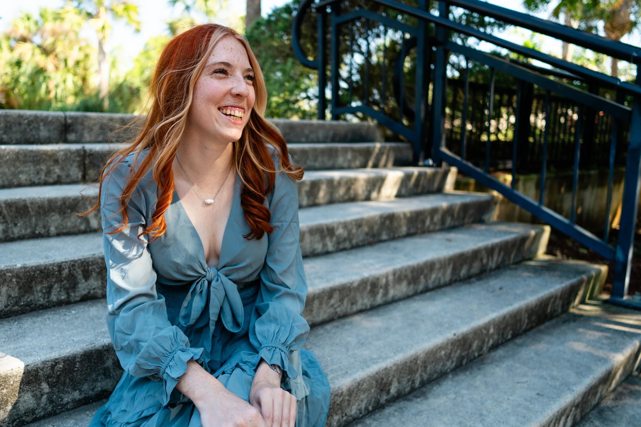 A smiling woman with red hair sits on outdoor stone stairs next to a black metal railing, surrounded by greenery.