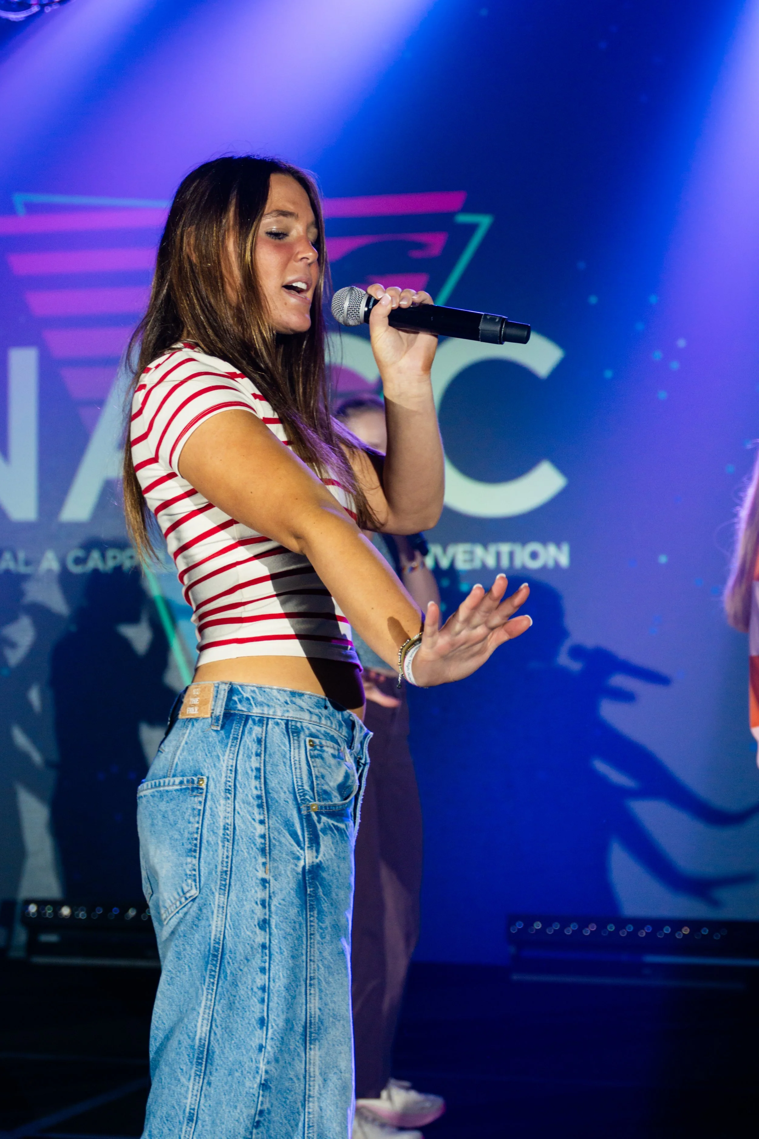 A woman with long brown hair, wearing a red and white striped crop top and high-waisted blue jeans, singing into a microphone on stage with a purple and blue background.