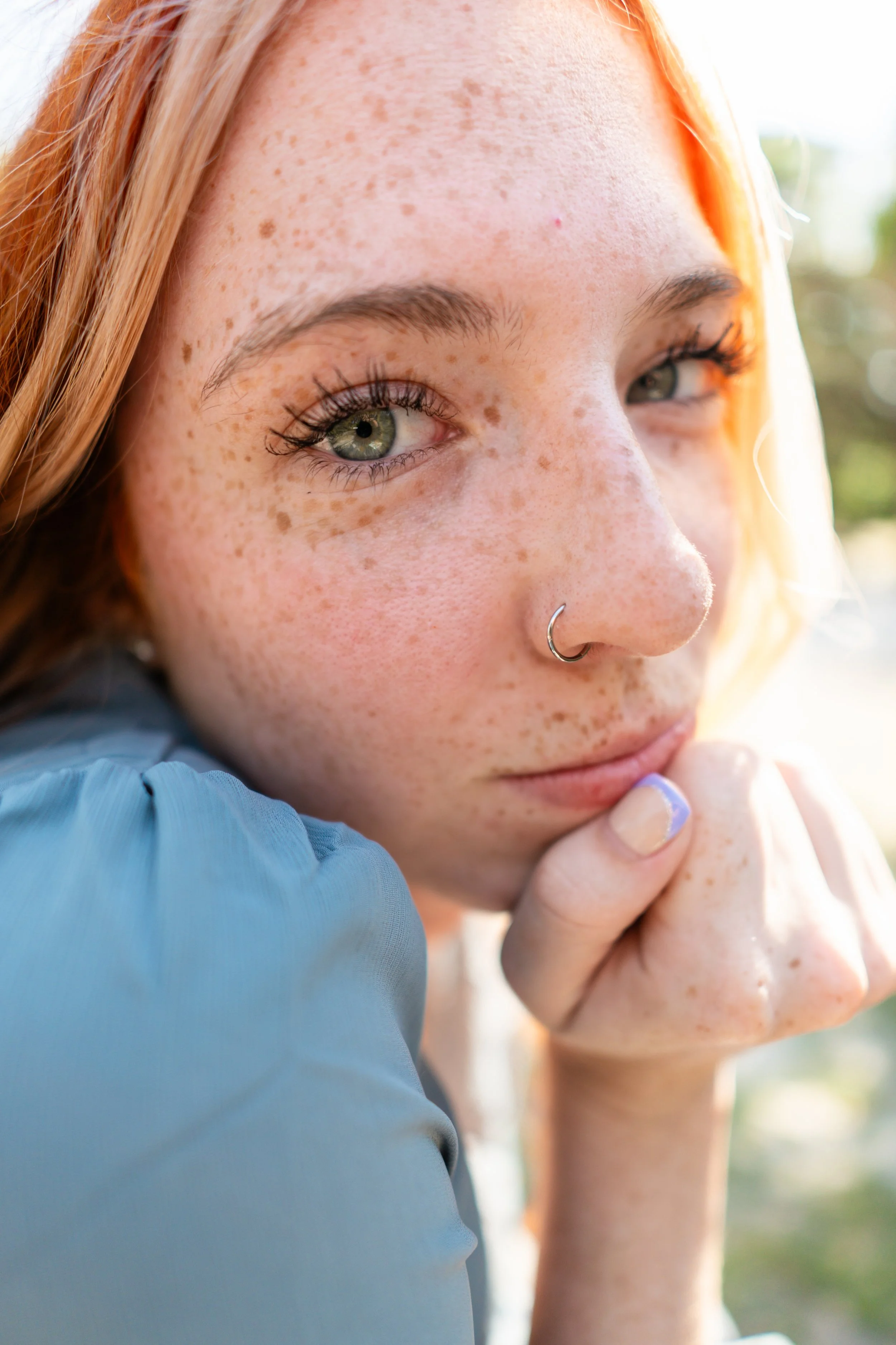 Close-up of a young woman with red hair, freckles, light green eyes, wearing a nose ring, and resting her chin on her hand.