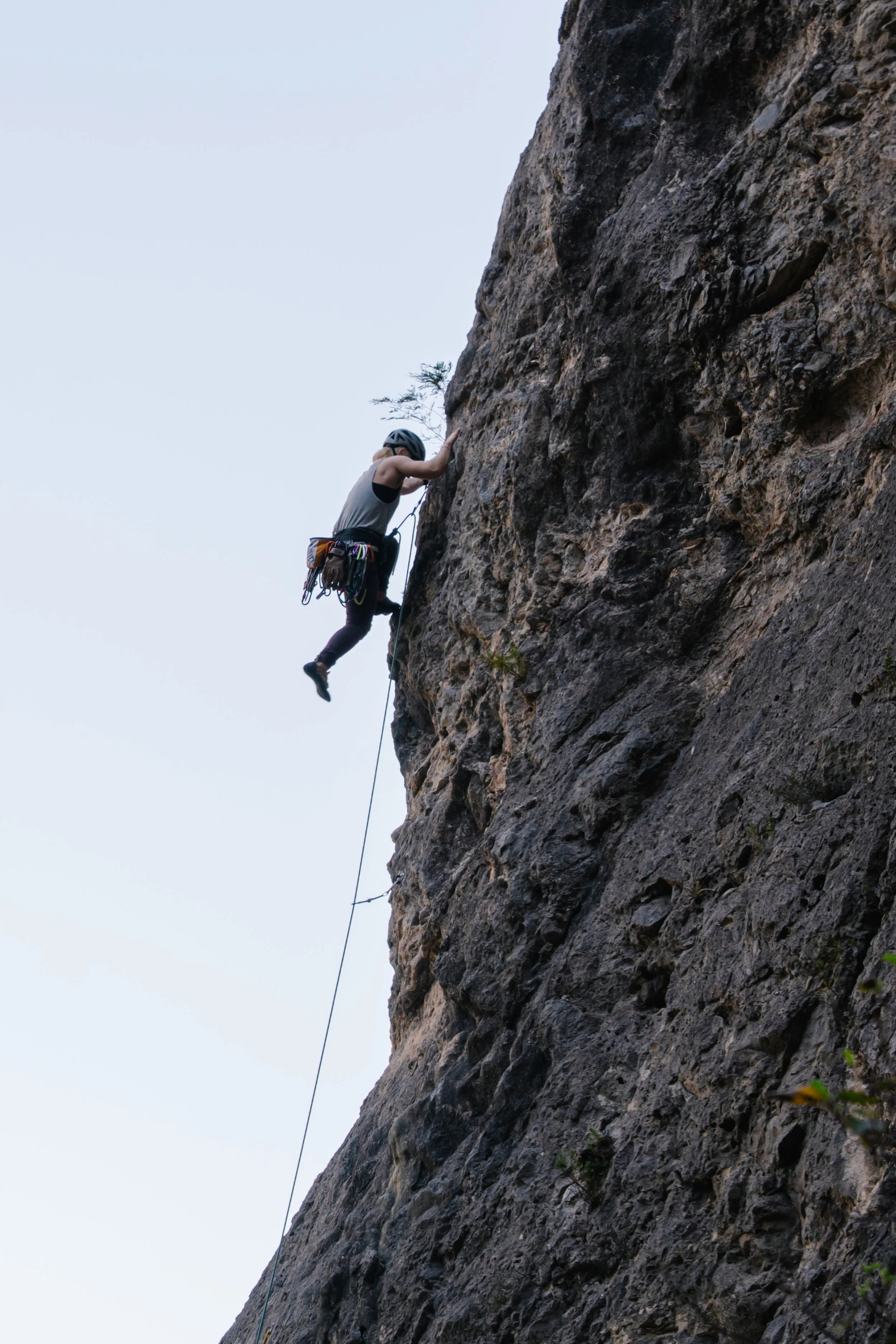 A person rock climbing on a steep cliff, wearing a helmet and harness, with climbing gear attached at their waist.