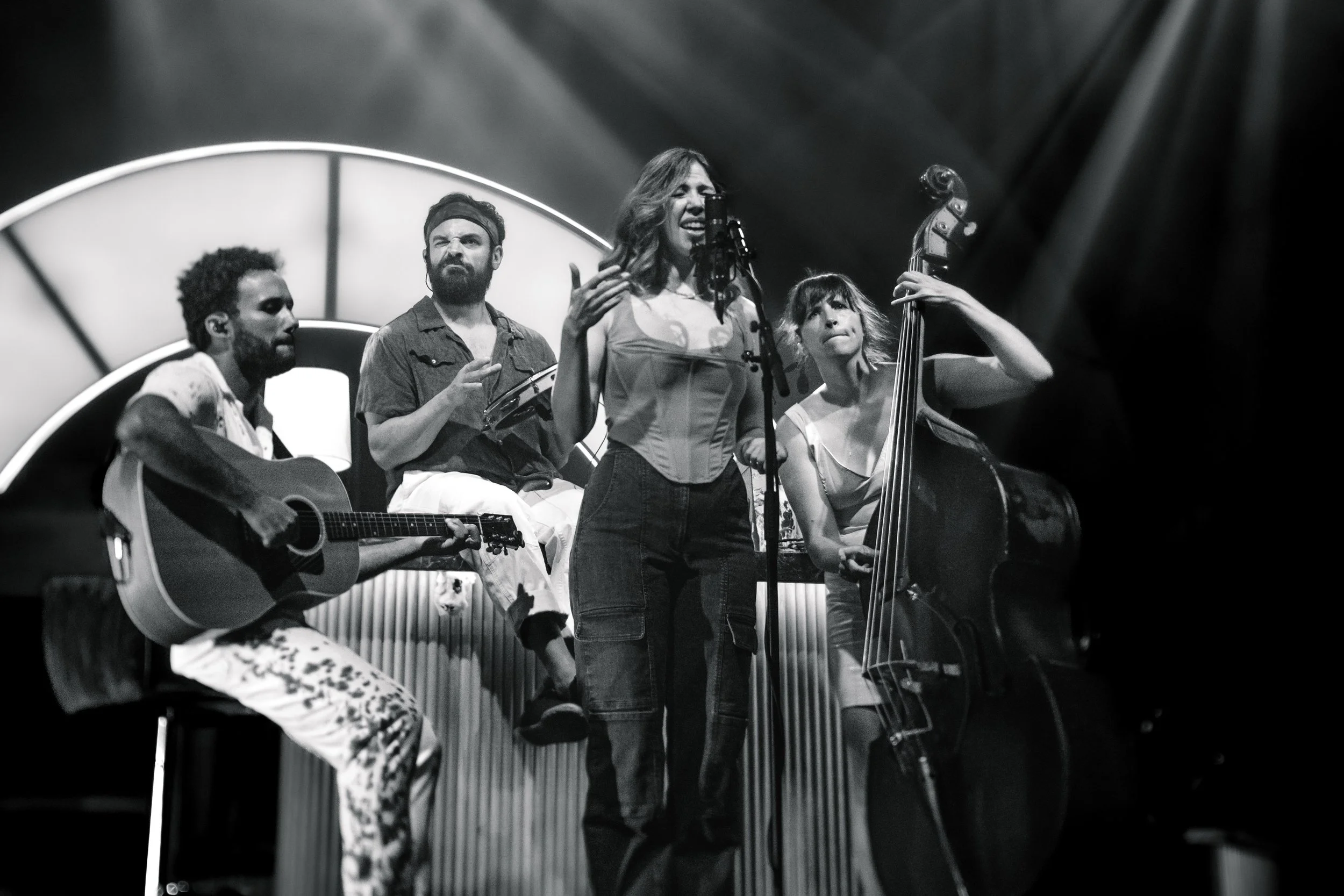 A black and white photo of four musicians performing on stage, with a woman singing into a microphone, a guitarist, a percussionist, and a bassist. The background features an illuminated circular design.