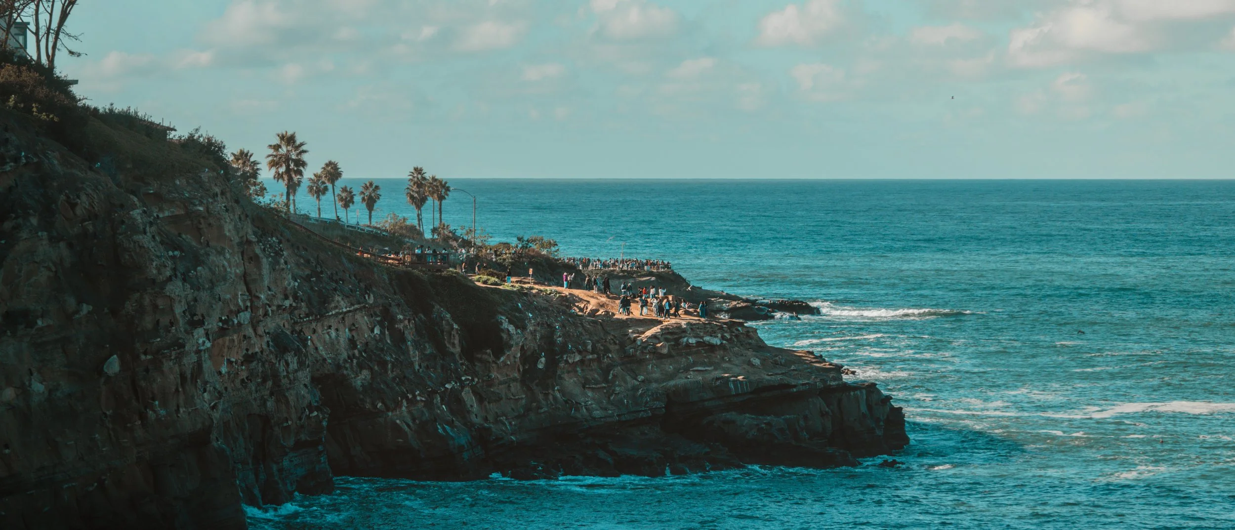 Cliffs along the coast with a crowd of people at the edge, palm trees in the background, ocean waves crashing below, and a partly cloudy sky.