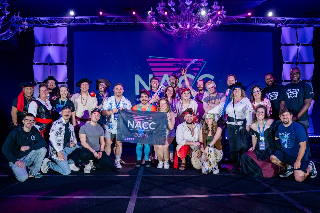 Group of people dressed in cosplay costumes and casual clothing on stage, holding a banner that reads 'NACC 2026', with a large screen behind them displaying the NACC logo and a starry background.
