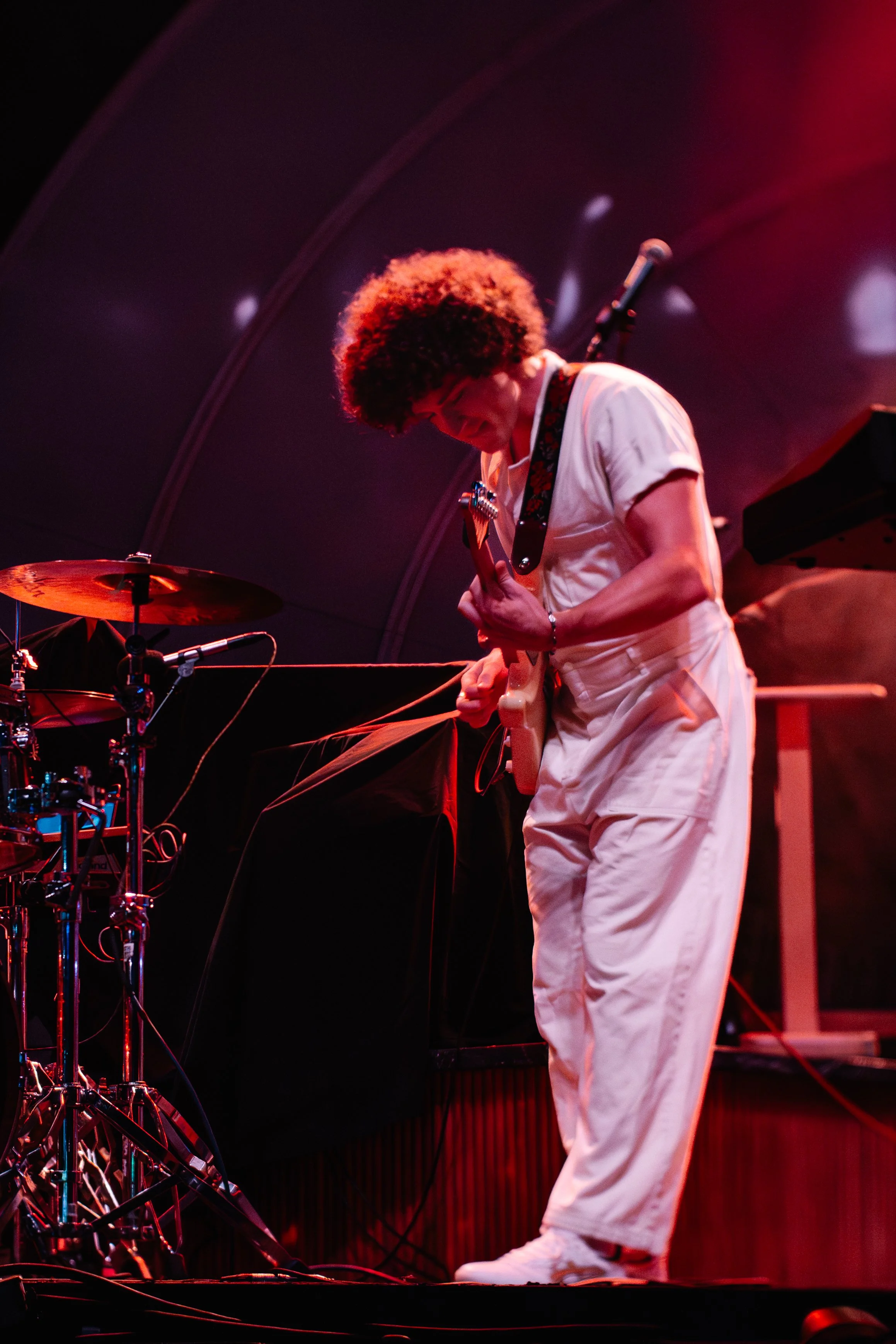 A musician wearing a white jumpsuit playing an electric guitar on stage, with drums and equipment visible in the background.