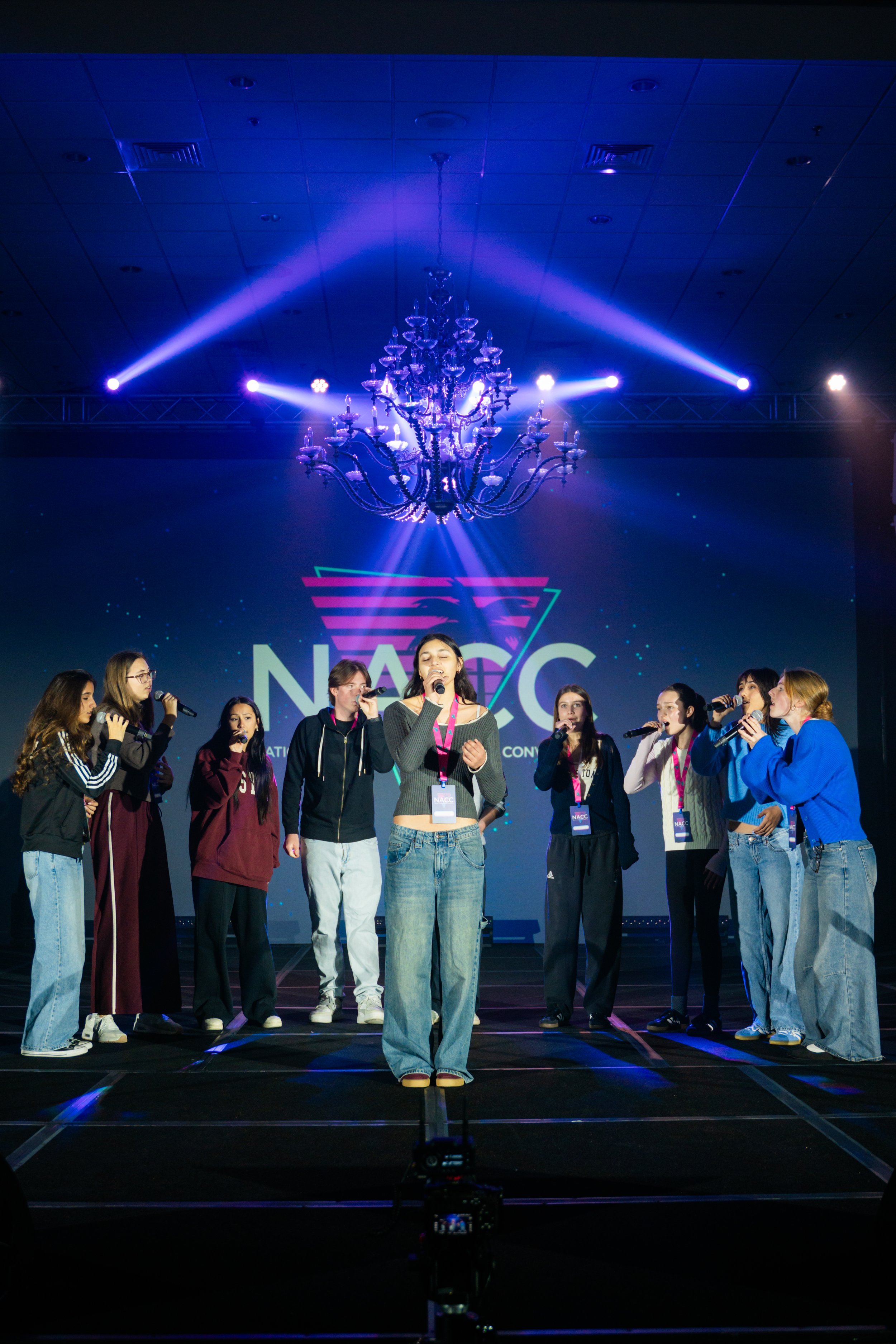 Group of young people on stage at a conference, with a large screen behind them displaying the logo "NACC" and a chandelier hanging from the ceiling, lit with vibrant purple and blue lights.
