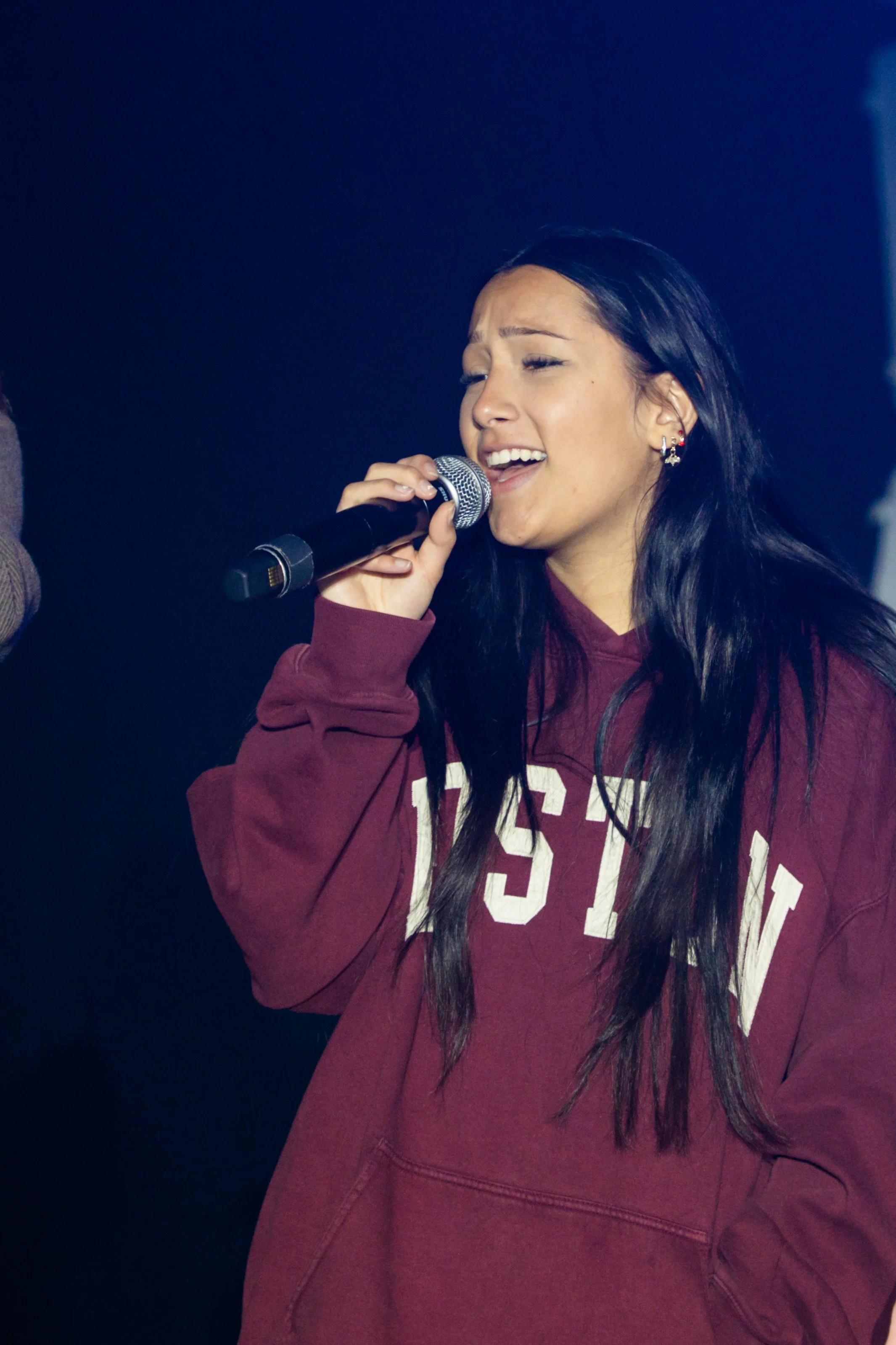 A young woman with long dark hair wearing a maroon sweatshirt singing into a microphone, with closed eyes and an expressive face.