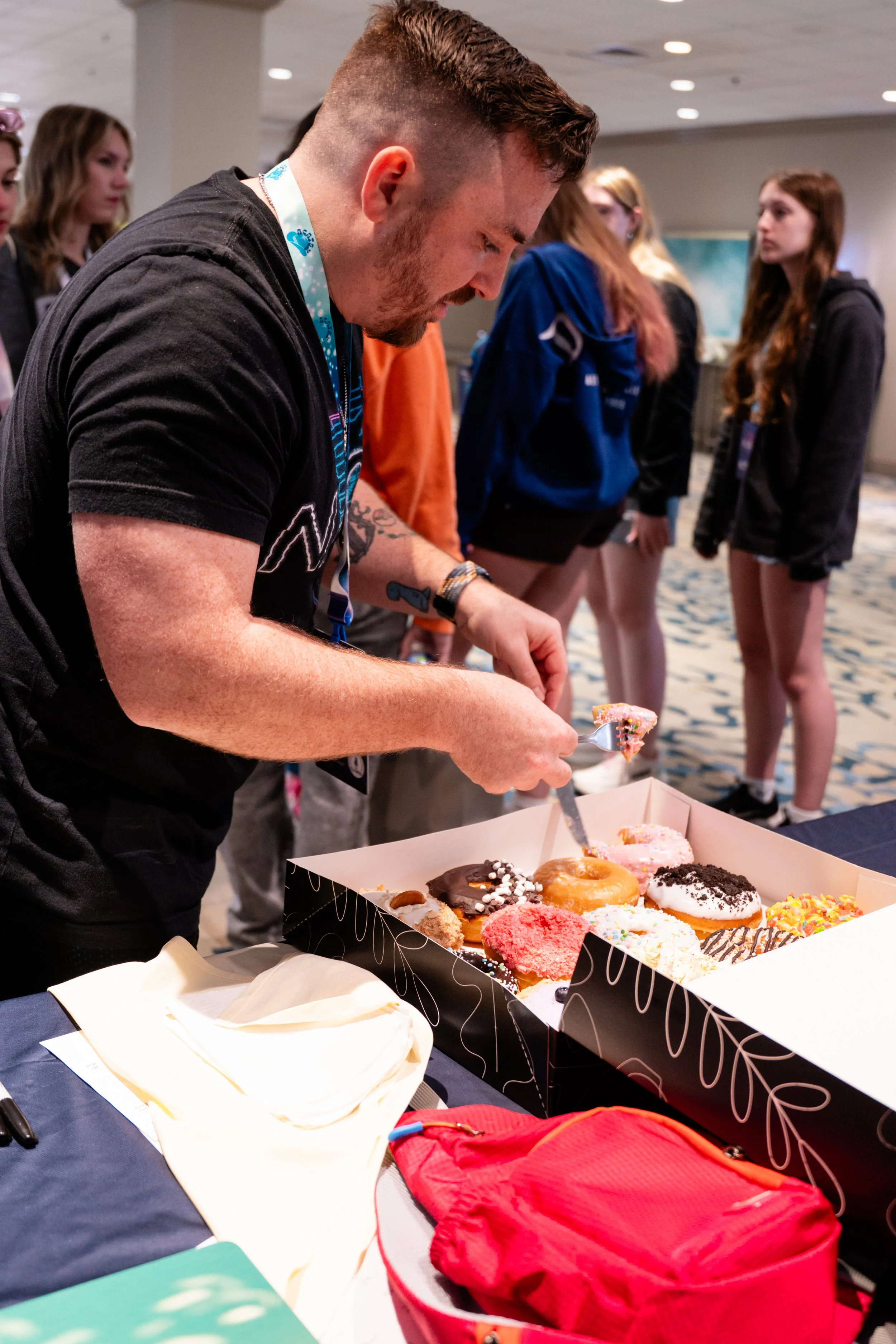 A man with a beard and tattoos on his arm is cutting and serving decorated donuts from a box on a table, with a group of young women in casual clothing in the background.