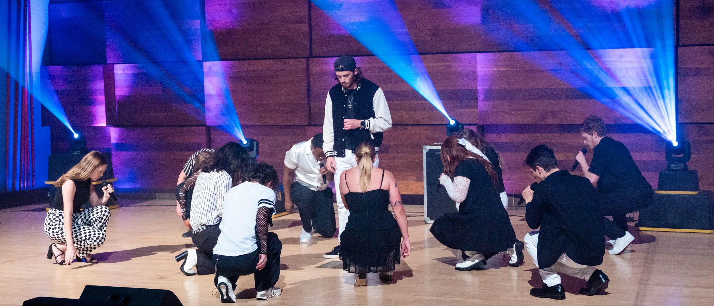 A group of young people are performing a dance routine on a stage with colorful lights. Some are kneeling, and one person is standing, holding a microphone.