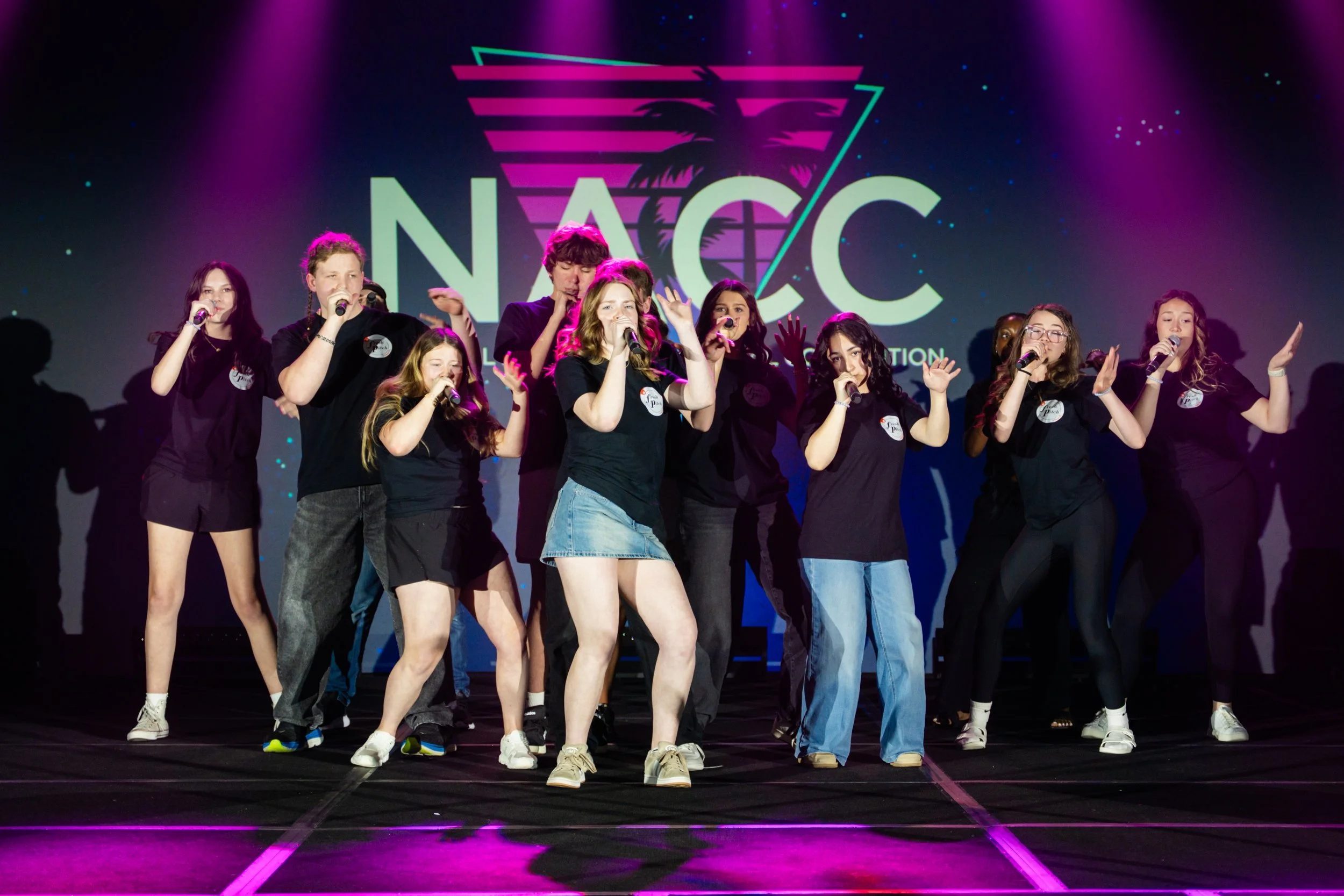Group of young people performing on stage with a large neon 'NACC' logo with a palm tree and colorful stripes in the background.