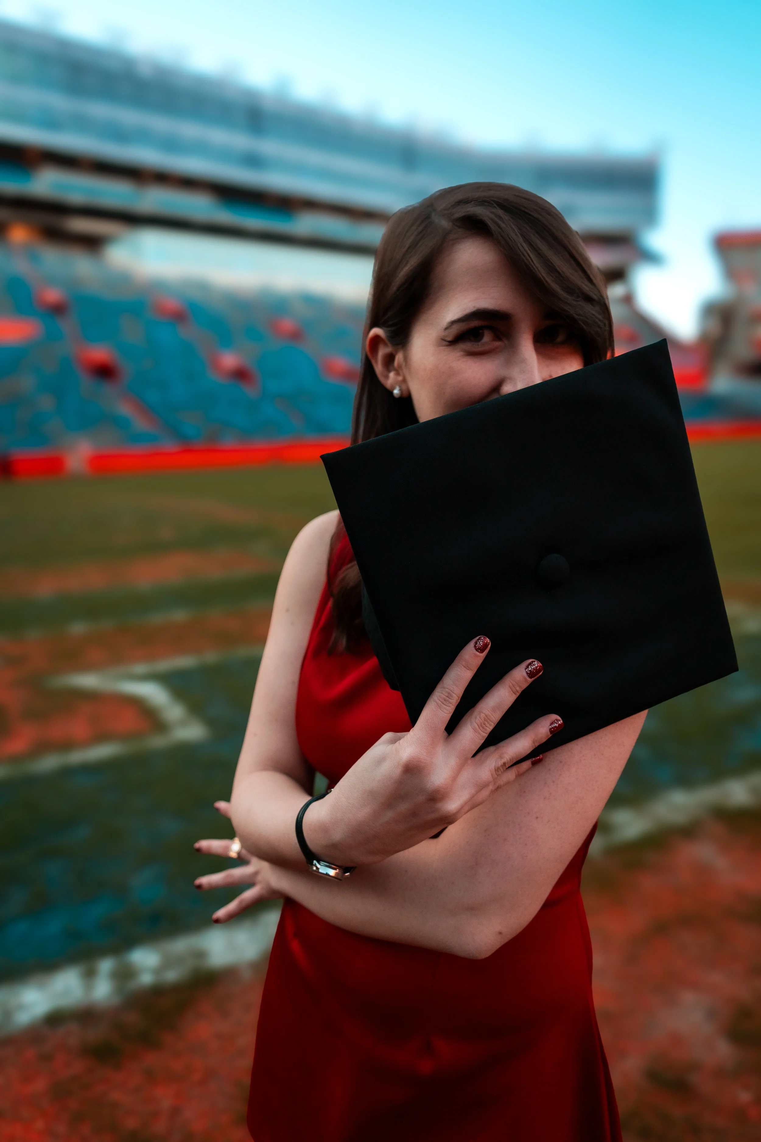 A woman in a red dress holding a graduation cap in front of her face on a sports field, with a stadium background.