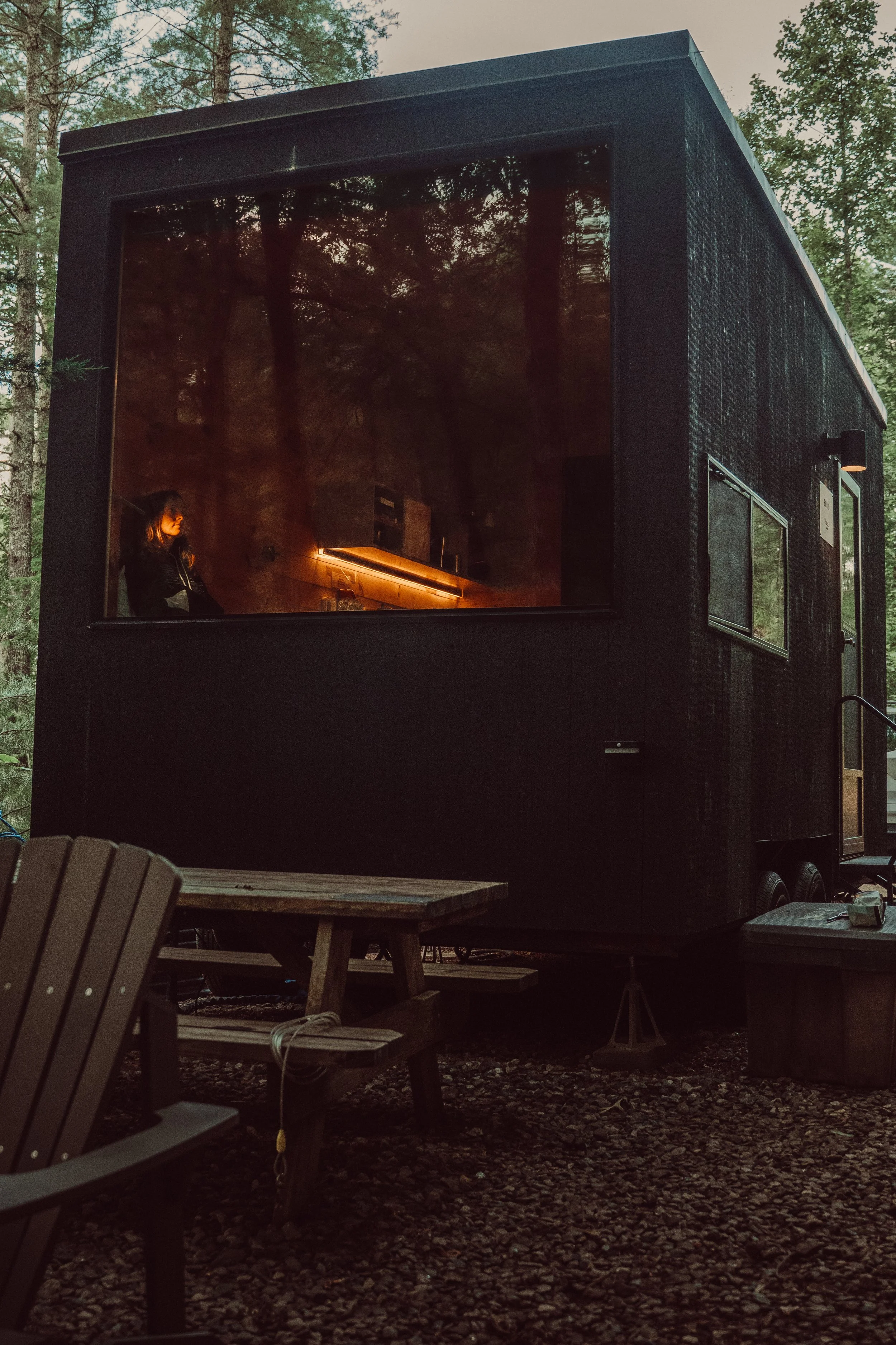 A black camper trailer parked outdoors in a wooded area at dusk, with a woman inside looking out the large front window.