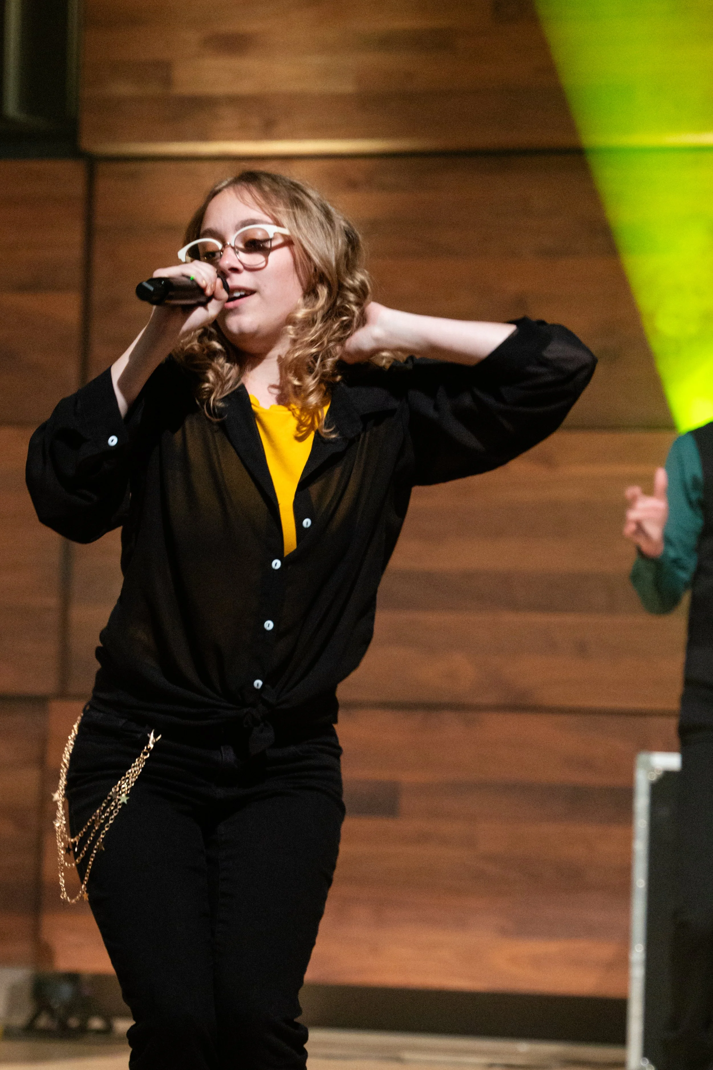 A young woman with curly blond hair and glasses singing into a microphone on stage, wearing a black shirt, yellow t-shirt, and black pants with a chain accessory.