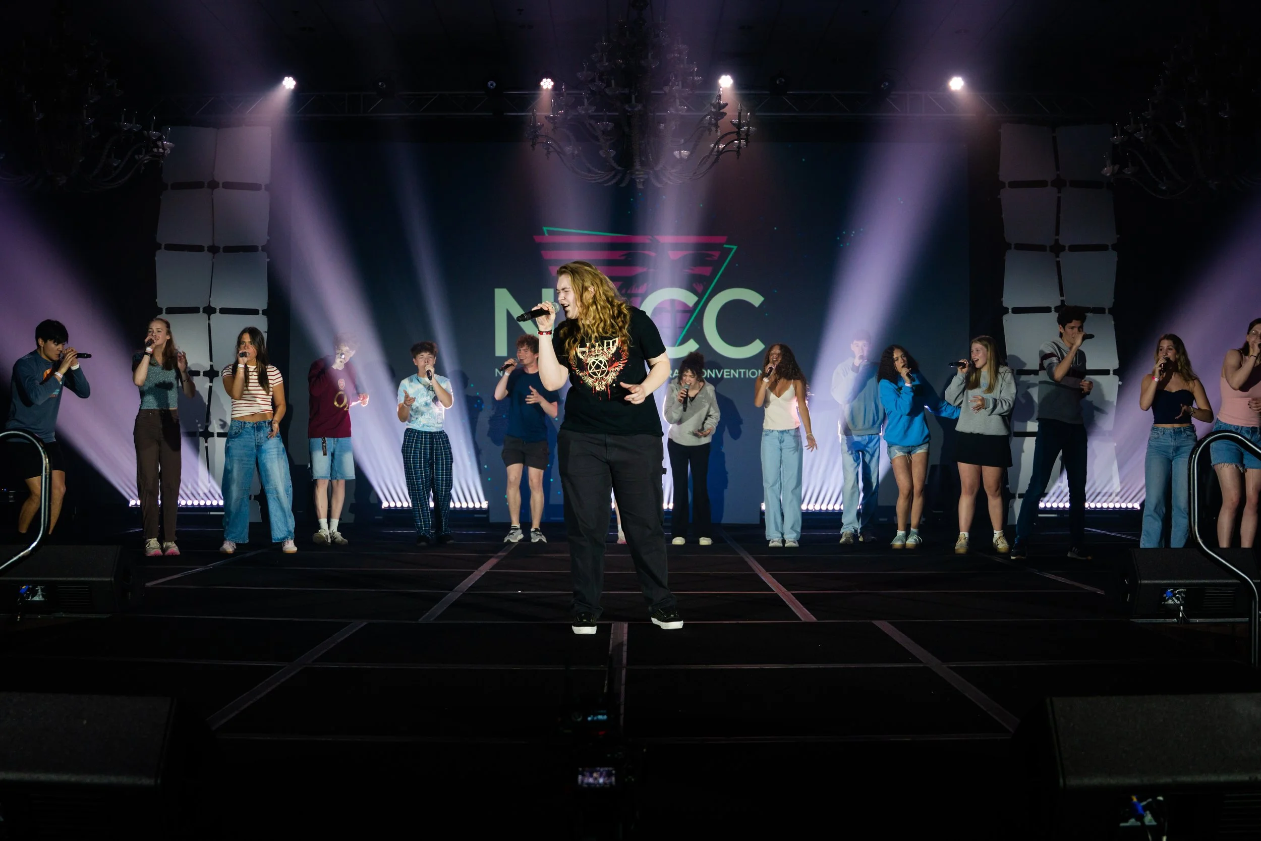 A group of young people performing on stage with a singer at the center and a large screen behind displaying a logo and the text 'NAC'. The stage has bright spotlights and a chandelier overhead.