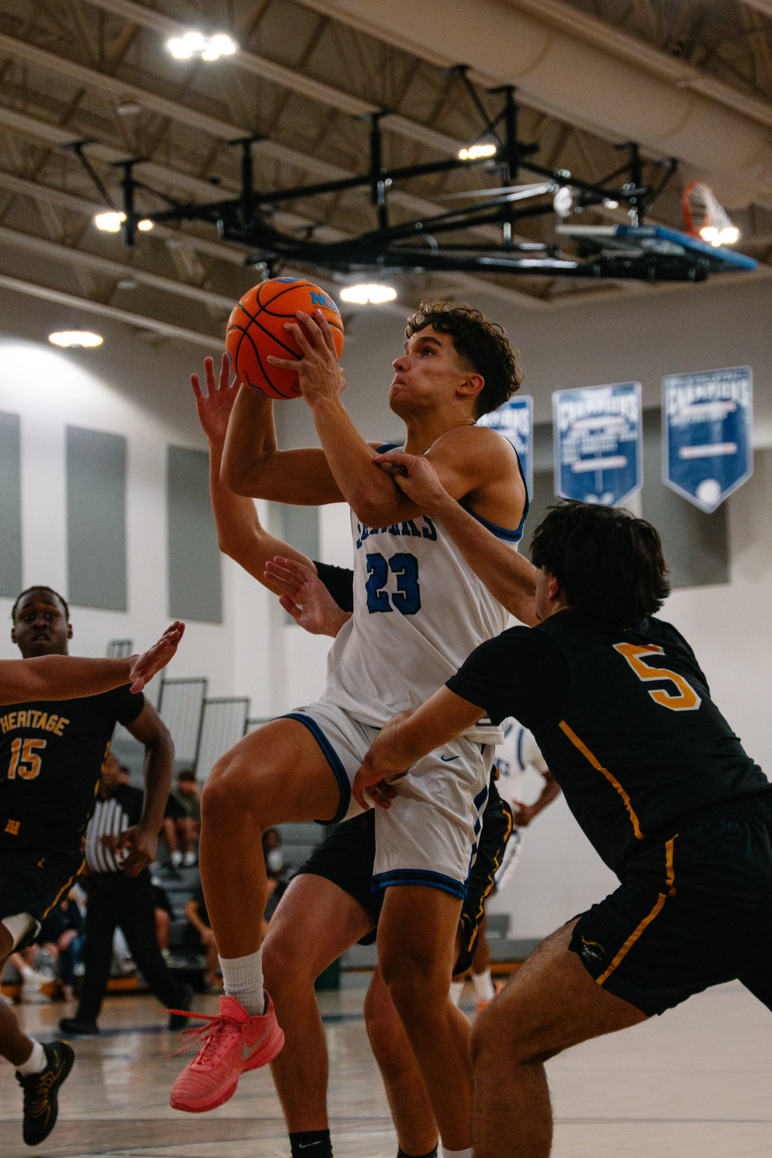 A basketball player wearing a white jersey with the number 23 is jumping to shoot or control the ball while being guarded closely by an opponent in a black jersey with the number 5. The scene takes place in an indoor basketball gym with banners hangi