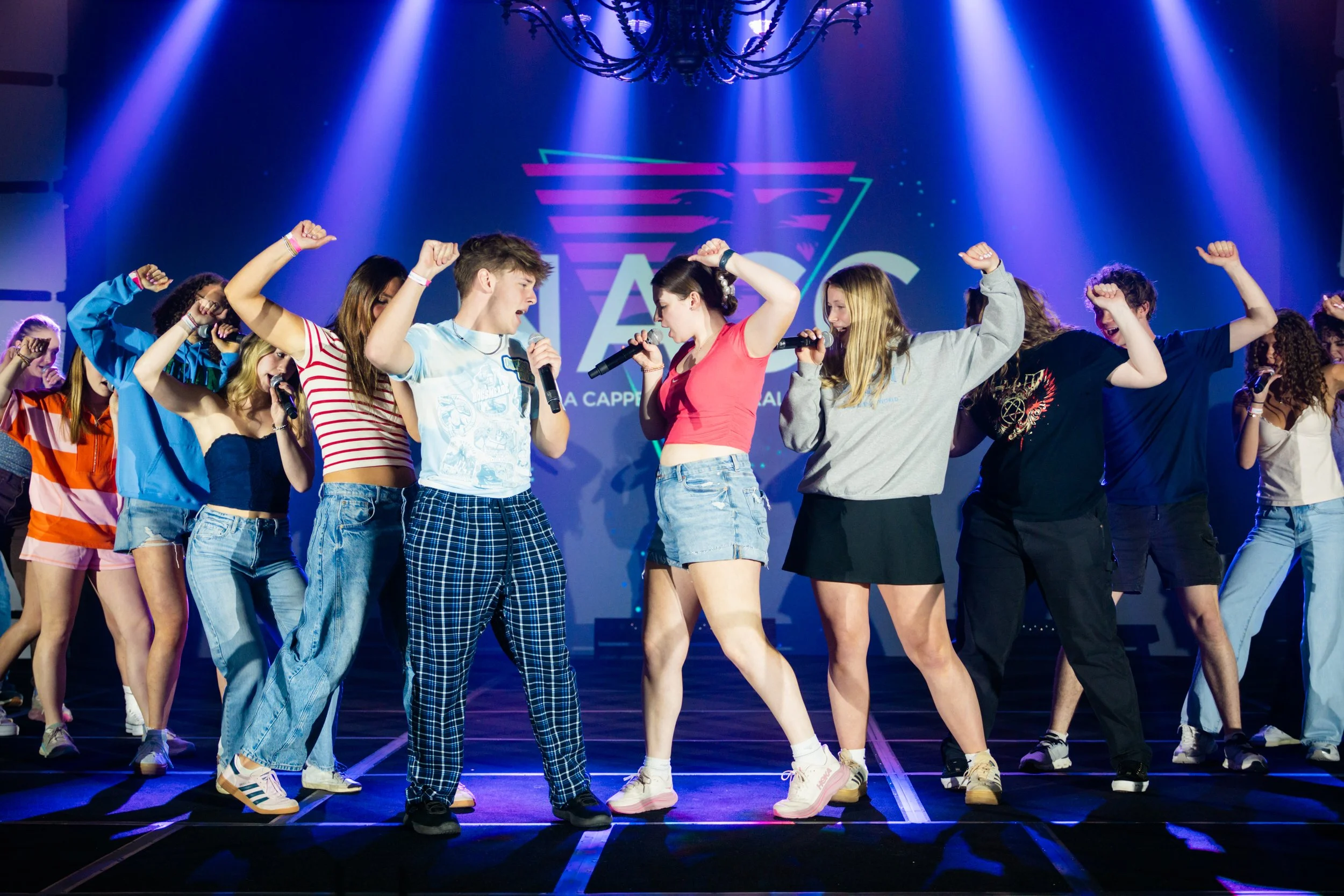 Group of young teenagers on stage performing together, some holding microphones, under colorful stage lighting.