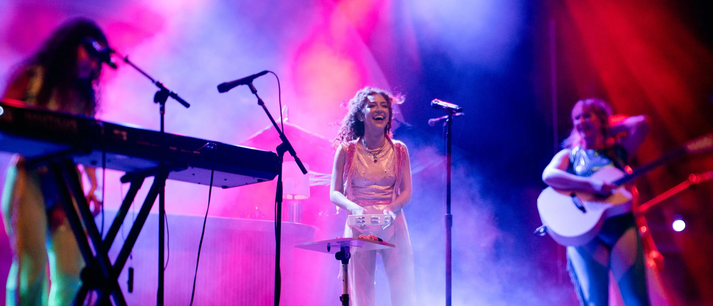 A group of female musicians performing on stage with colorful lighting and fog effects, including one woman singing and playing electronic instruments, another playing guitar, and another on keyboard.