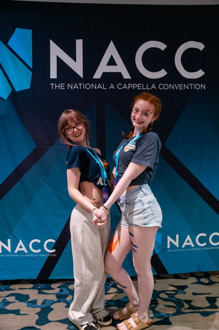 Two young women holding hands and smiling at the camera in front of a blue NACC banner at the National A Cappella Convention. They are wearing casual clothes and have face paint.