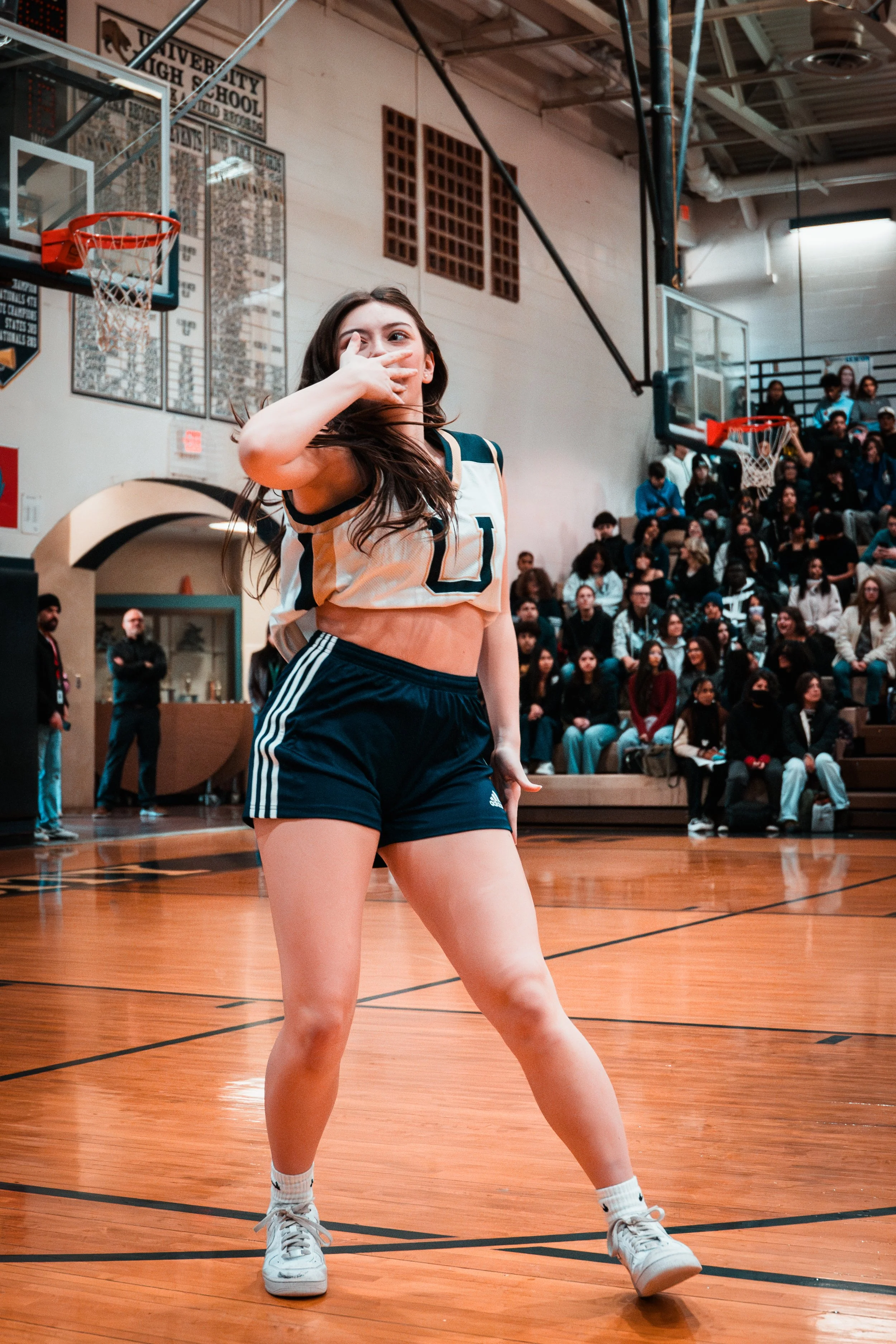 A female basketball player in a cream and navy uniform on a basketball court, striking a confident pose with her hand near her face, with a crowd seated in the bleachers behind her.