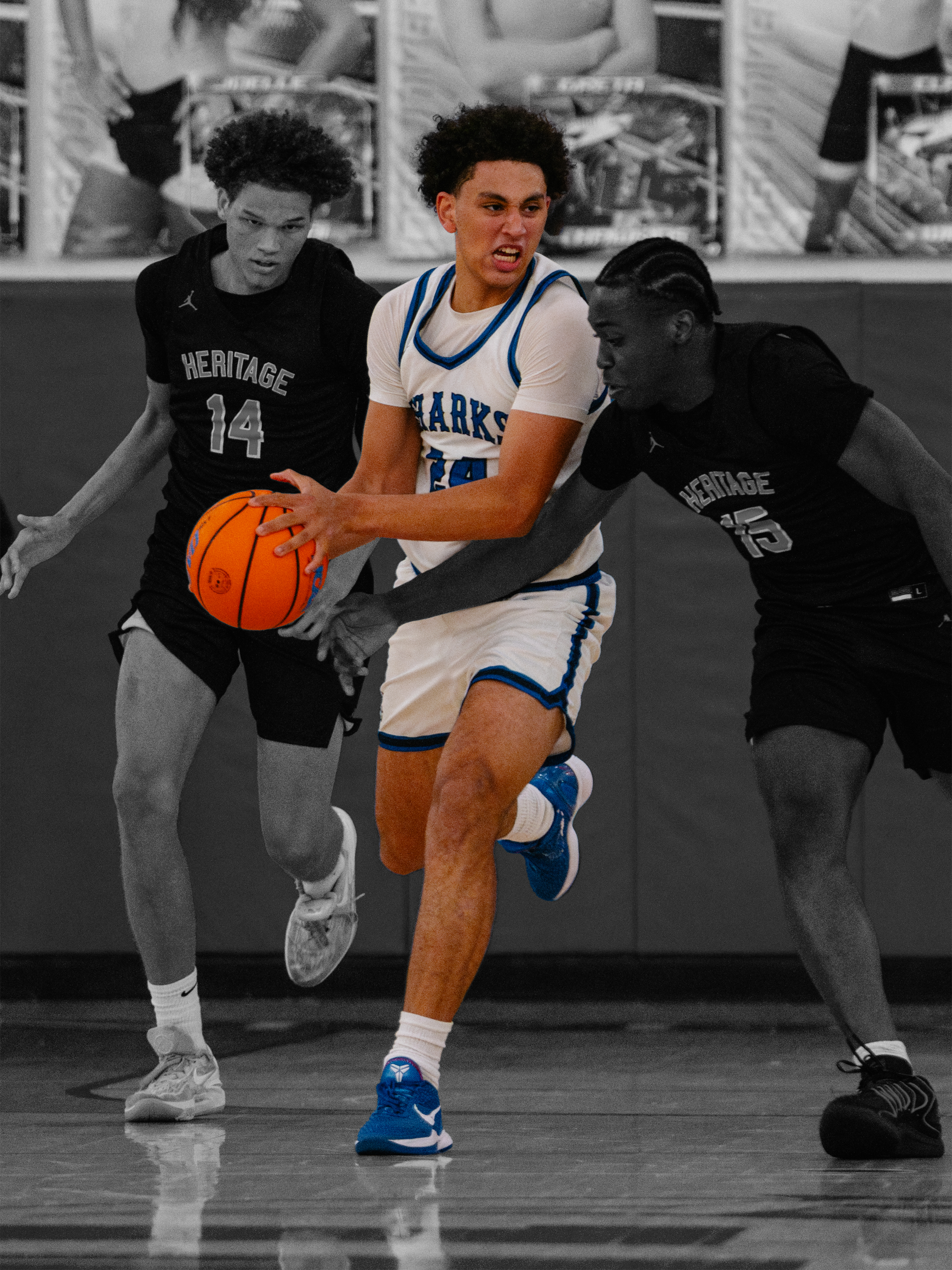 Two players from Heritage school, wearing black jerseys, are defending against a player in a white and blue jersey with 'SHARKS' on it, who is dribbling a basketball and trying to get past them during a game.