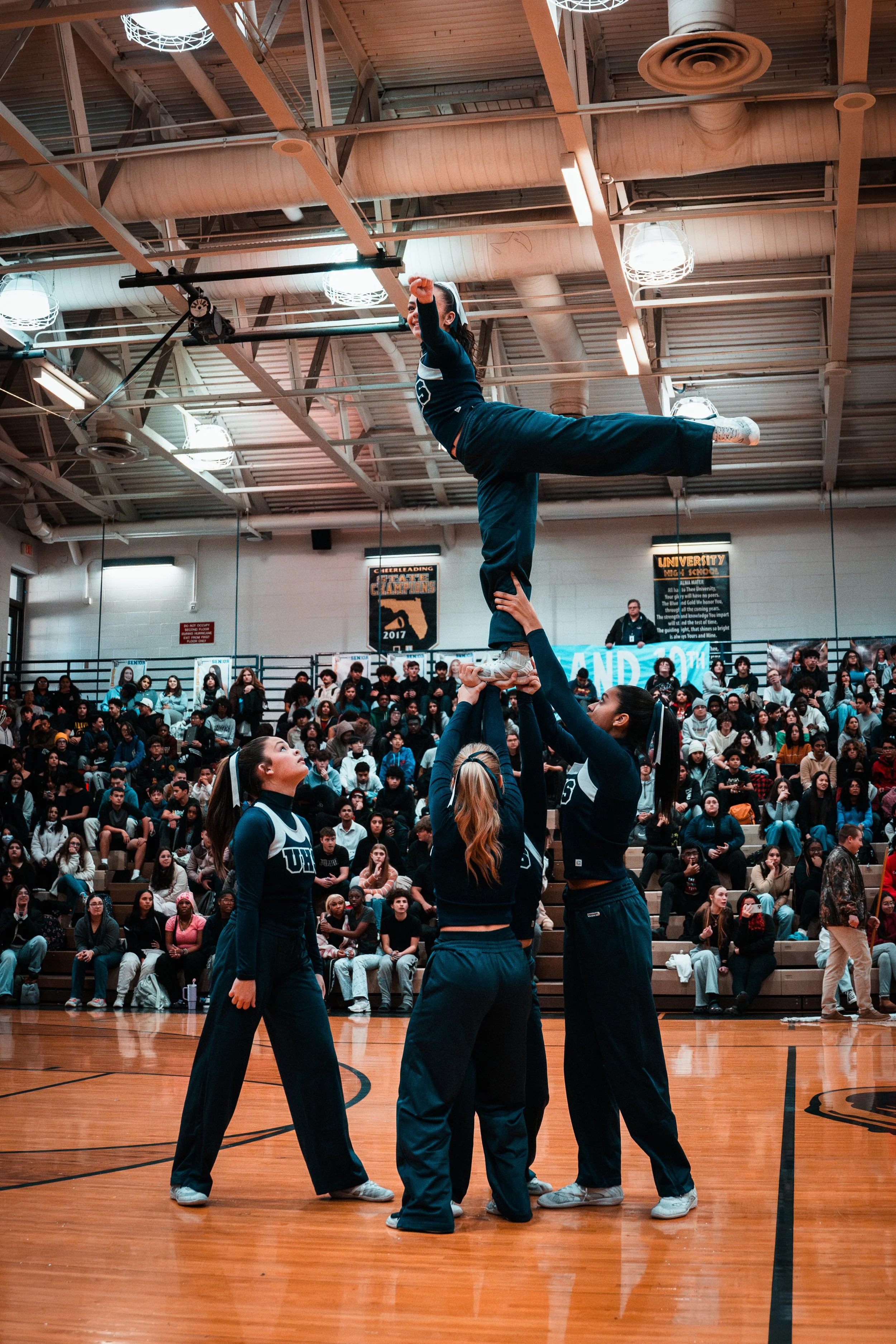 Cheerleaders perform a stunt in a gymnasium during a competition with a large audience watching.