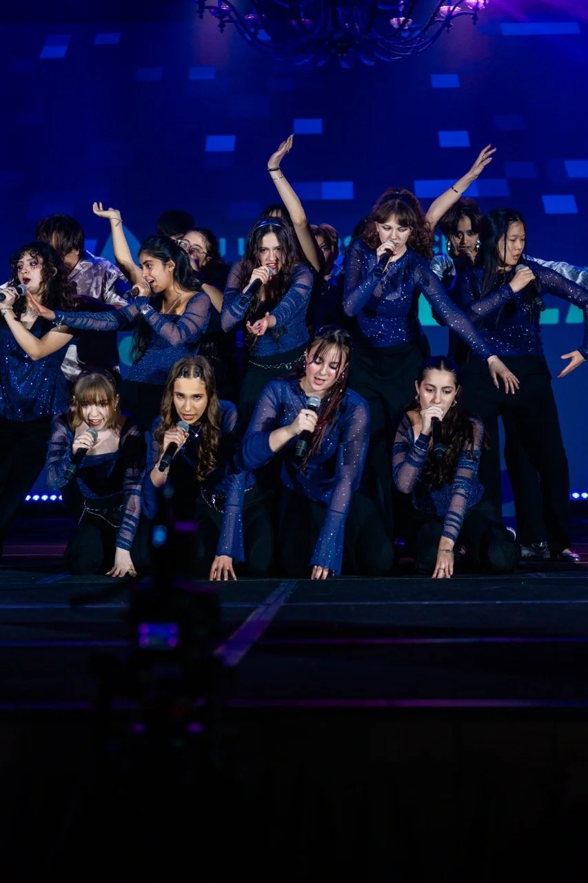 Group of teenage girls performing on stage with microphones, wearing blue sparkly costumes, under blue stage lighting.