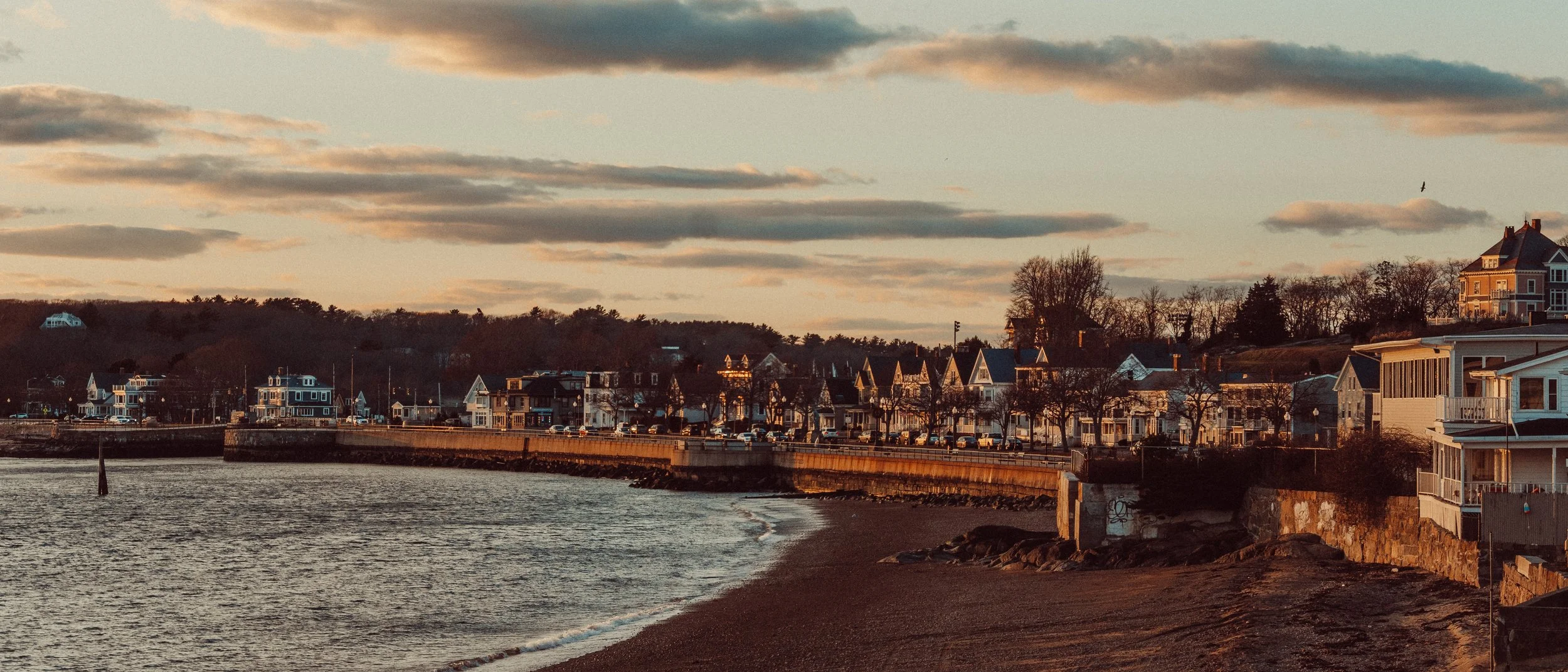 Sunset over a coastal town with houses and a waterfront promenade, with calm water and cloudy sky.