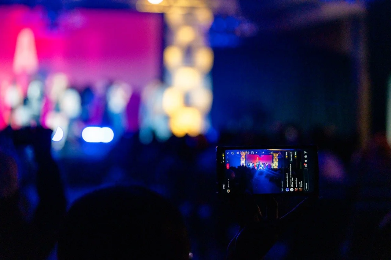 A concert or event stage with colorful lighting, seen in the background, and a person recording with a smartphone in the foreground.
