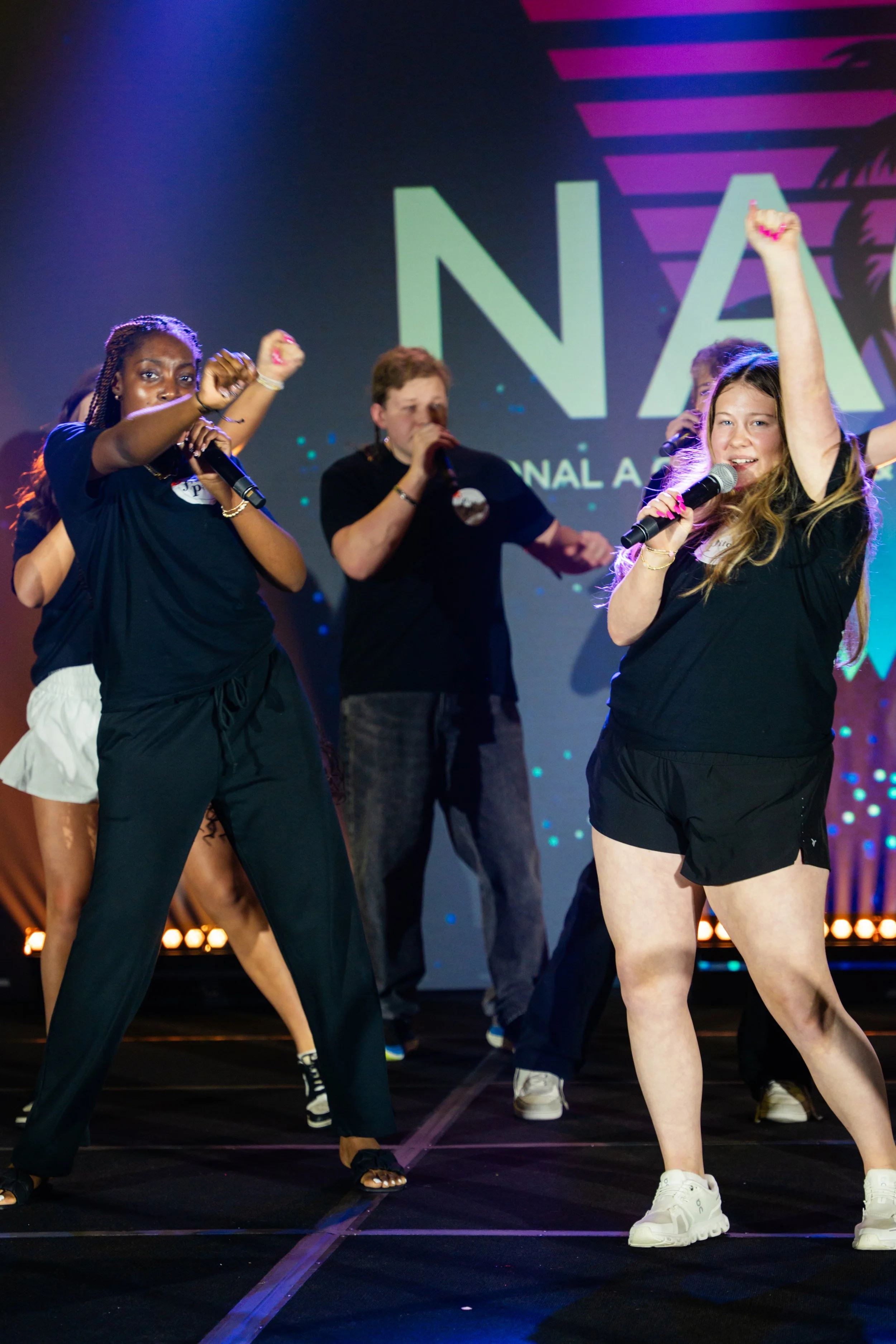 Group of young women and men dancing on stage at an event, with a man holding a microphone and a large screen displaying the word 'NA' in the background.
