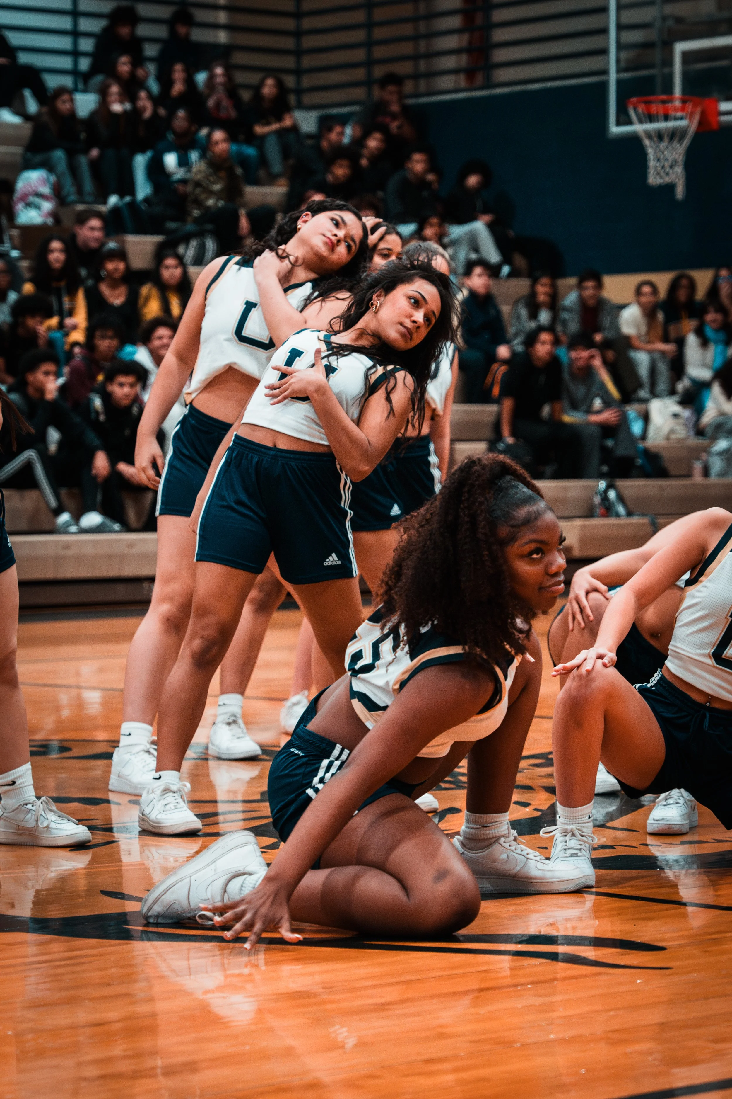 A group of female cheerleaders in matching sports uniforms performing a dance routine on a basketball court, with an audience seated in the background.