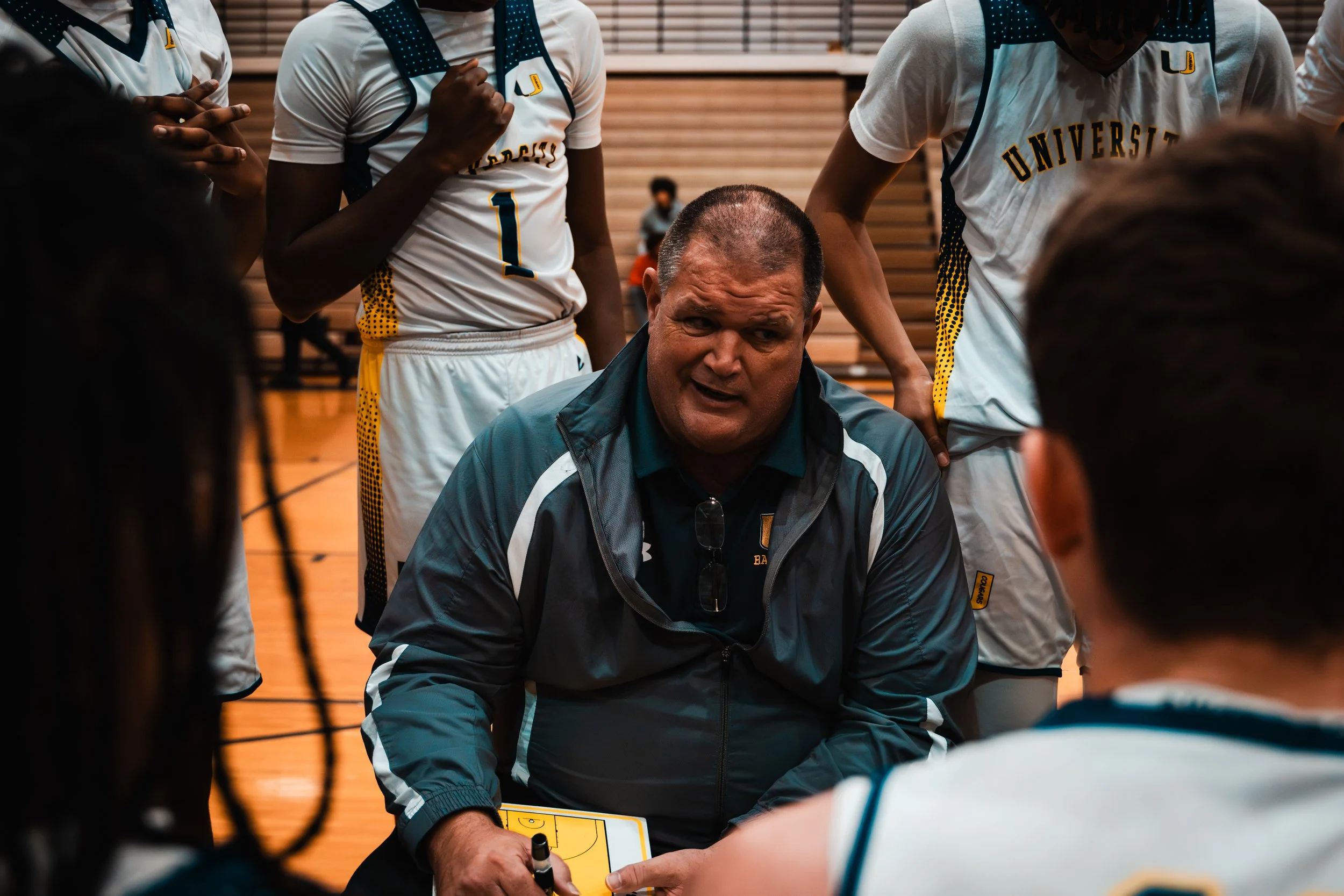 A basketball coach speaking to his team during a timeout in a gymnasium. The coach is crouched down, holding a marker, and surrounded by players in white and yellow uniforms with the word 'UNIVERSITY' on them.