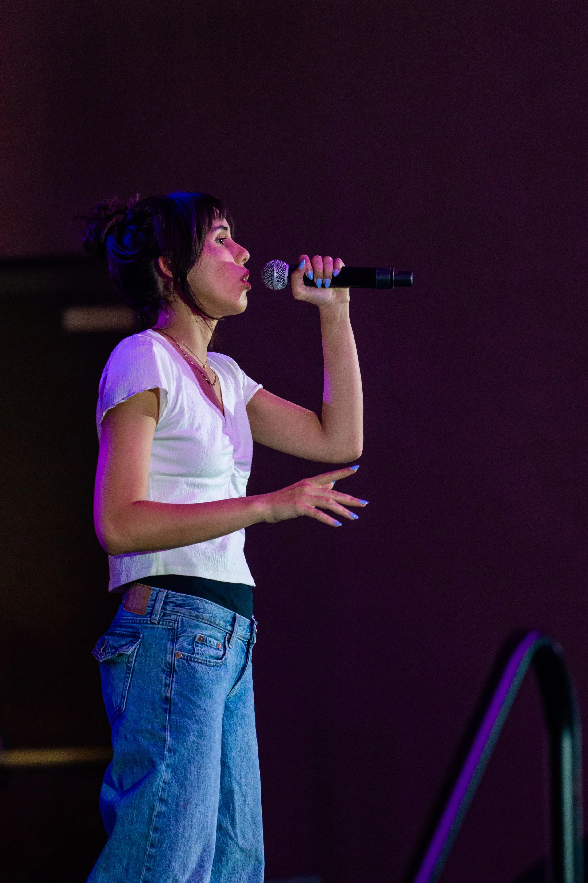 A woman with dark hair tied in a messy bun, wearing a white top and jeans, singing into a microphone on a dark stage with purple lighting.