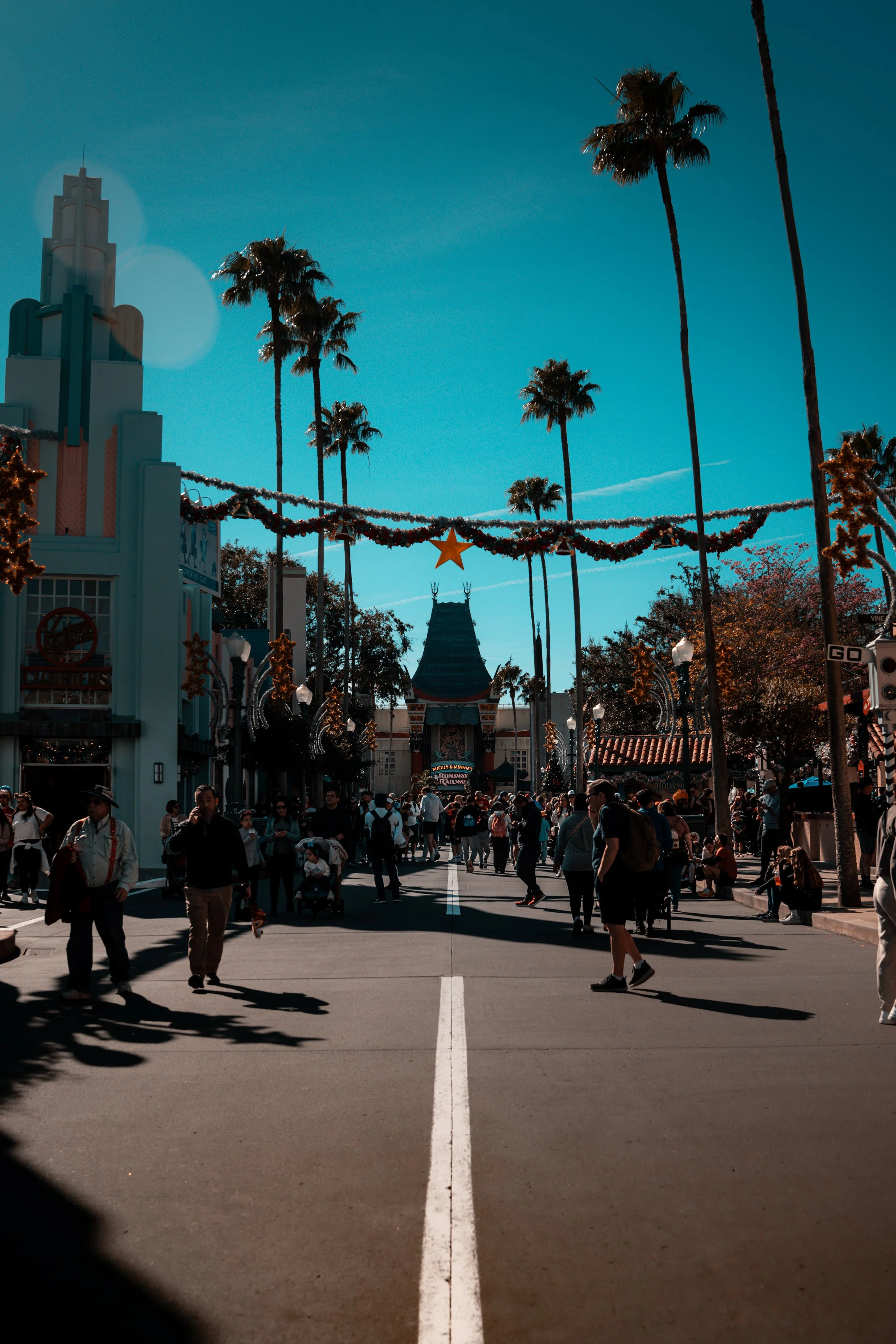 People walking in a themed amusement park with palm trees and decorative holiday banners, featuring a stylized building and a clock tower in the background.