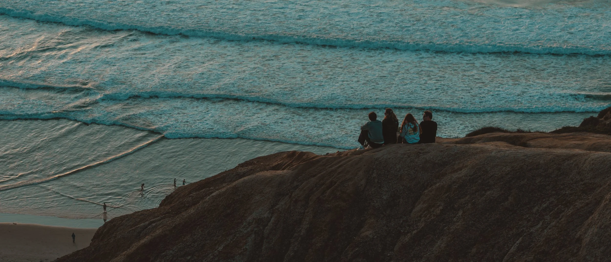 Four people sit on a rocky cliff overlooking the ocean at sunset, with waves rolling in below.