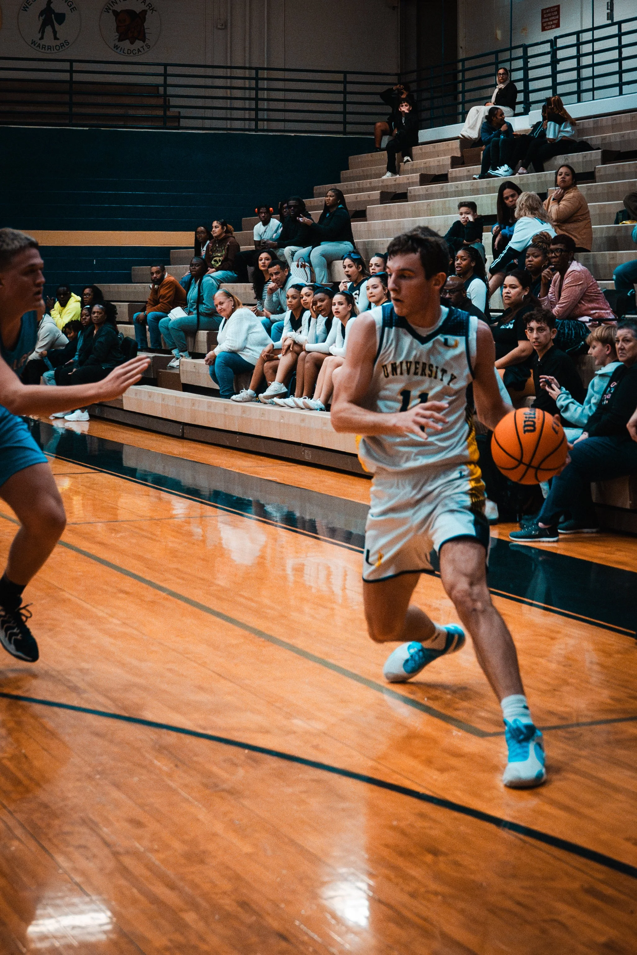 A basketball player in a white uniform dribbles the ball on the court, with spectators seated on the bleachers watching the game.