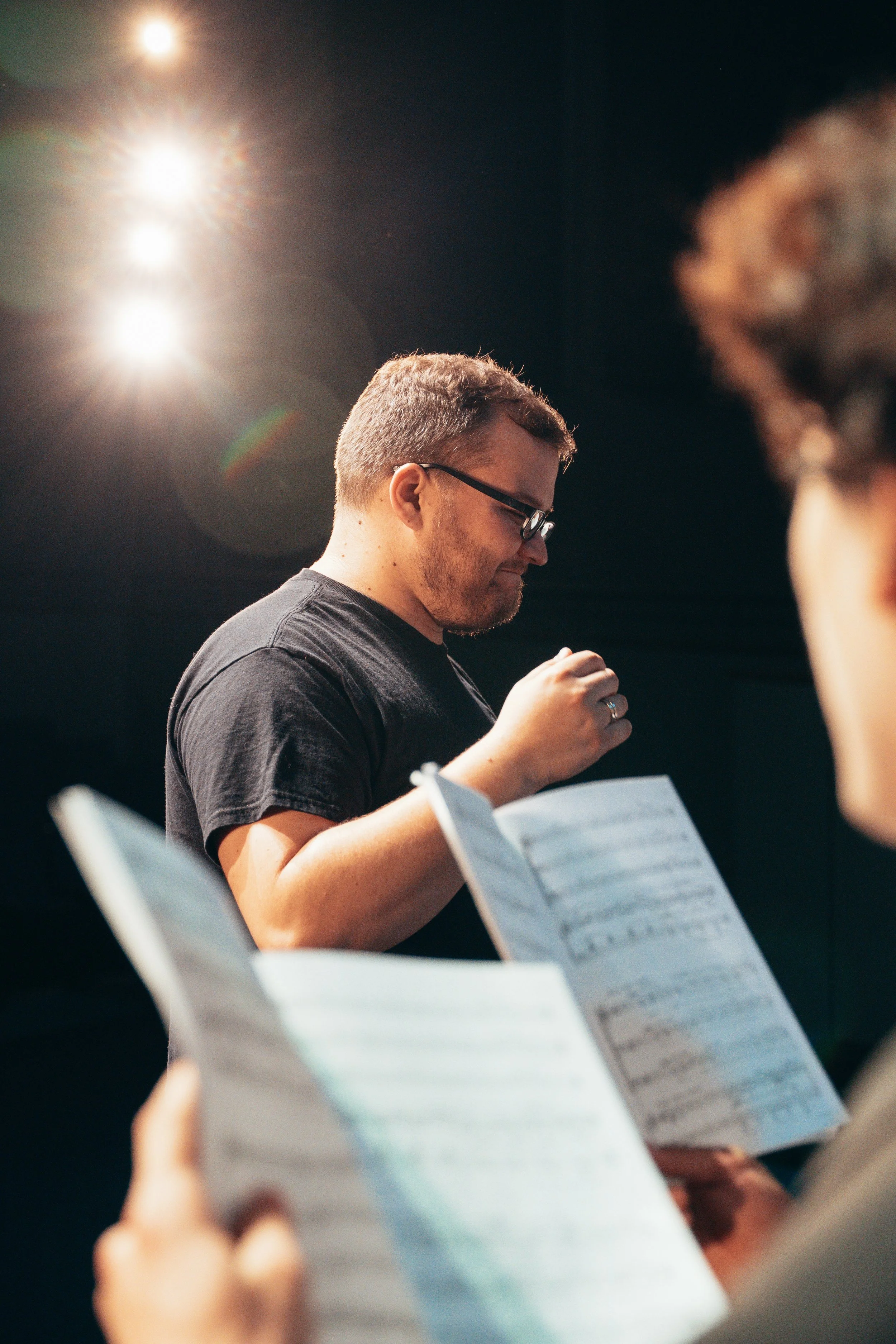 A man wearing glasses and a black t-shirt is holding a sheet of music or notes, with another person in front also holding a similar sheet. The scene appears to be in a rehearsal or practice space with bright stage lights overhead.