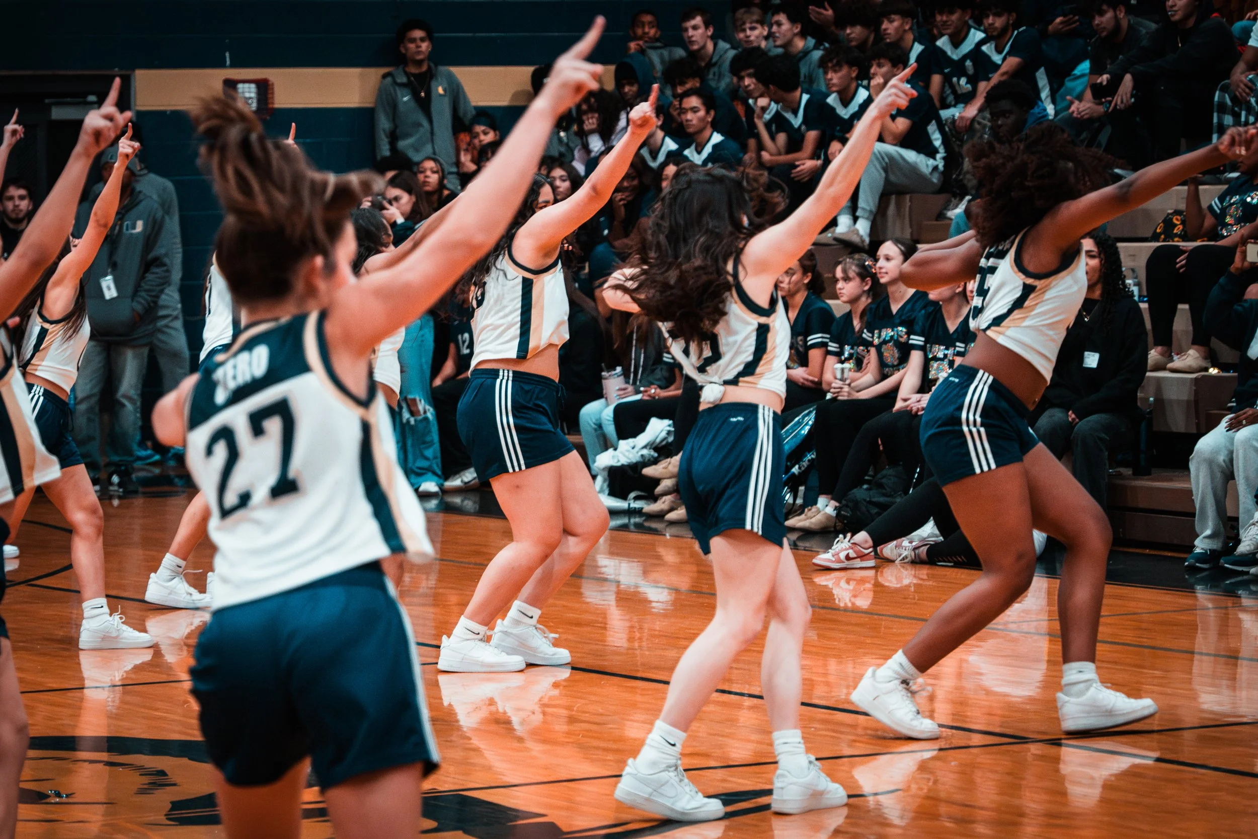 A group of young women cheerleaders performing a dance routine during a basketball game, with an audience in the background.