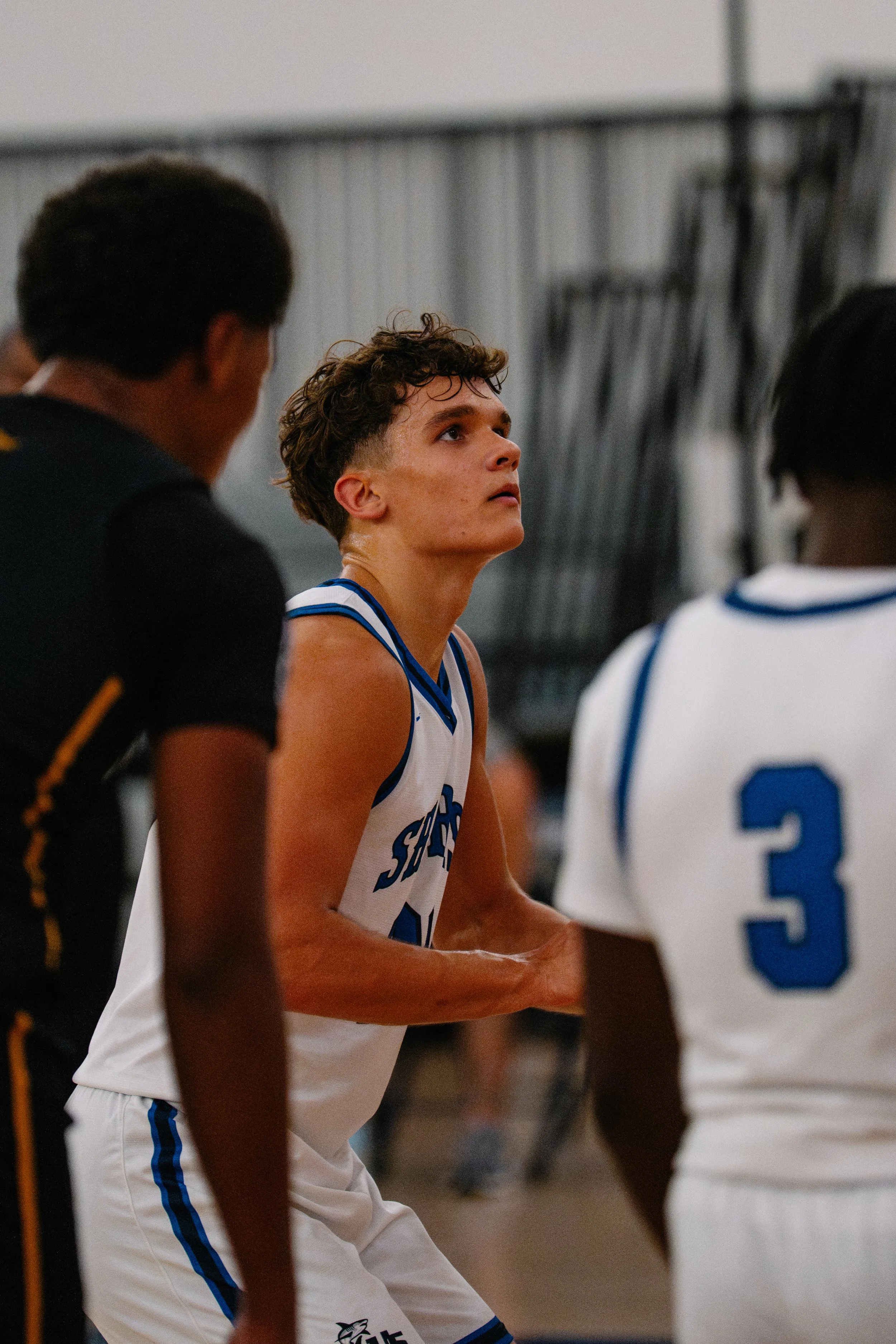 A young basketball player in a white uniform looks attentively during a game, with his hands clasped, surrounded by teammates and opponents in a gymnasium.