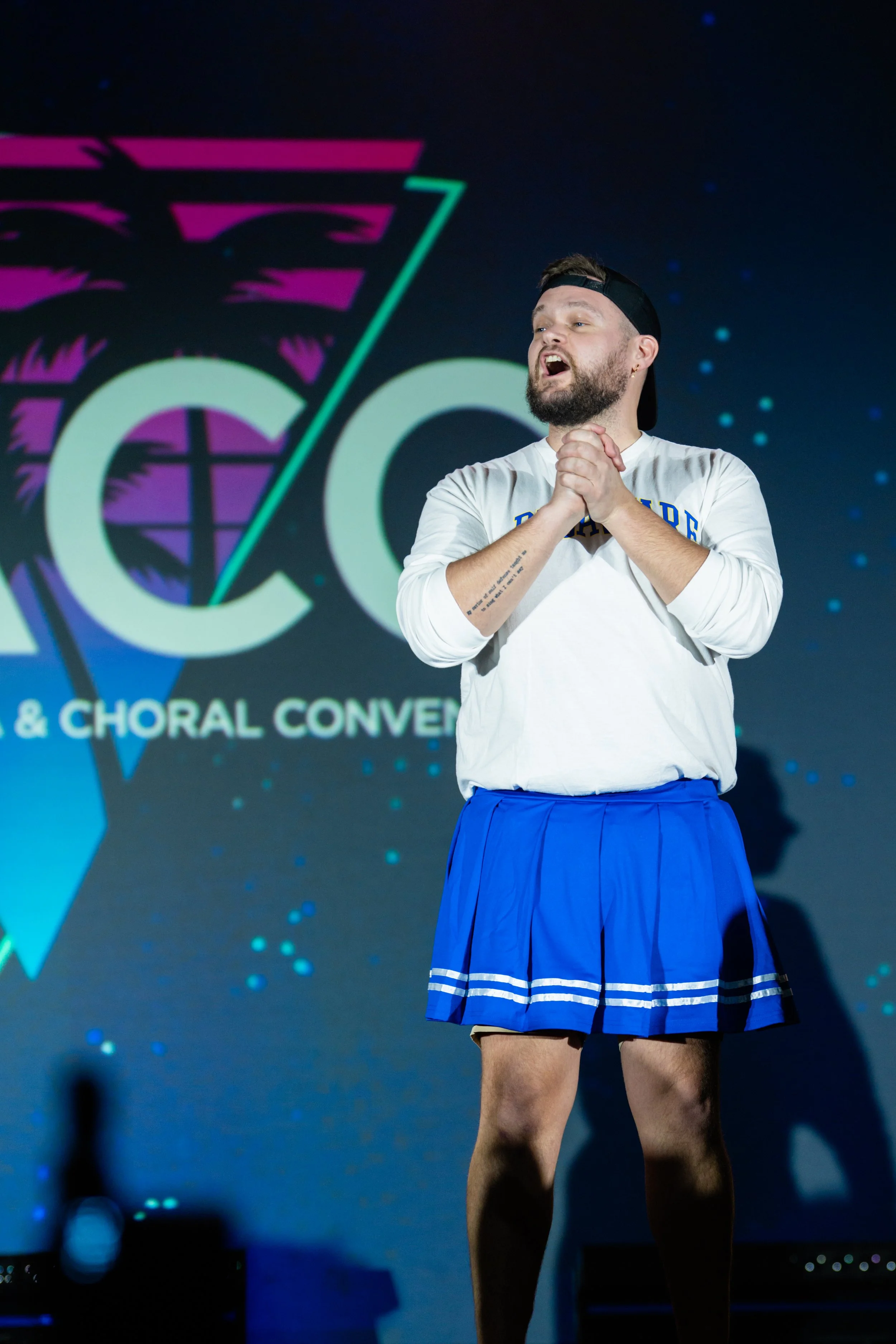 A man with a beard and a backward black cap is on stage, singing or speaking with his eyes closed and hands clasped. He is wearing a white shirt with writing and a blue pleated skirt, with a tattoo on his right forearm. Behind him is a dark backgroun
