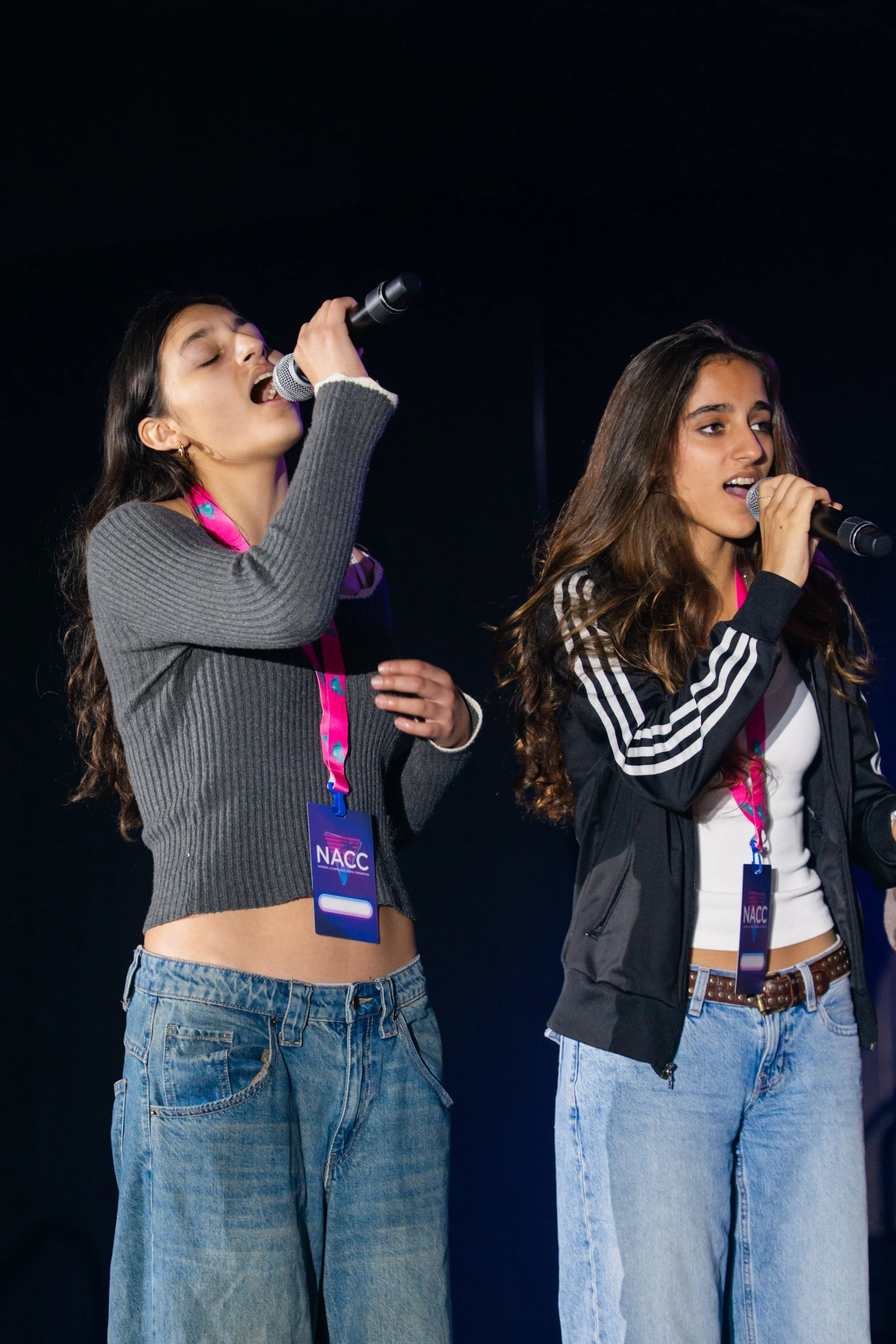 Two young women singing into microphones on stage, wearing casual clothing and passing badges that say NACC.