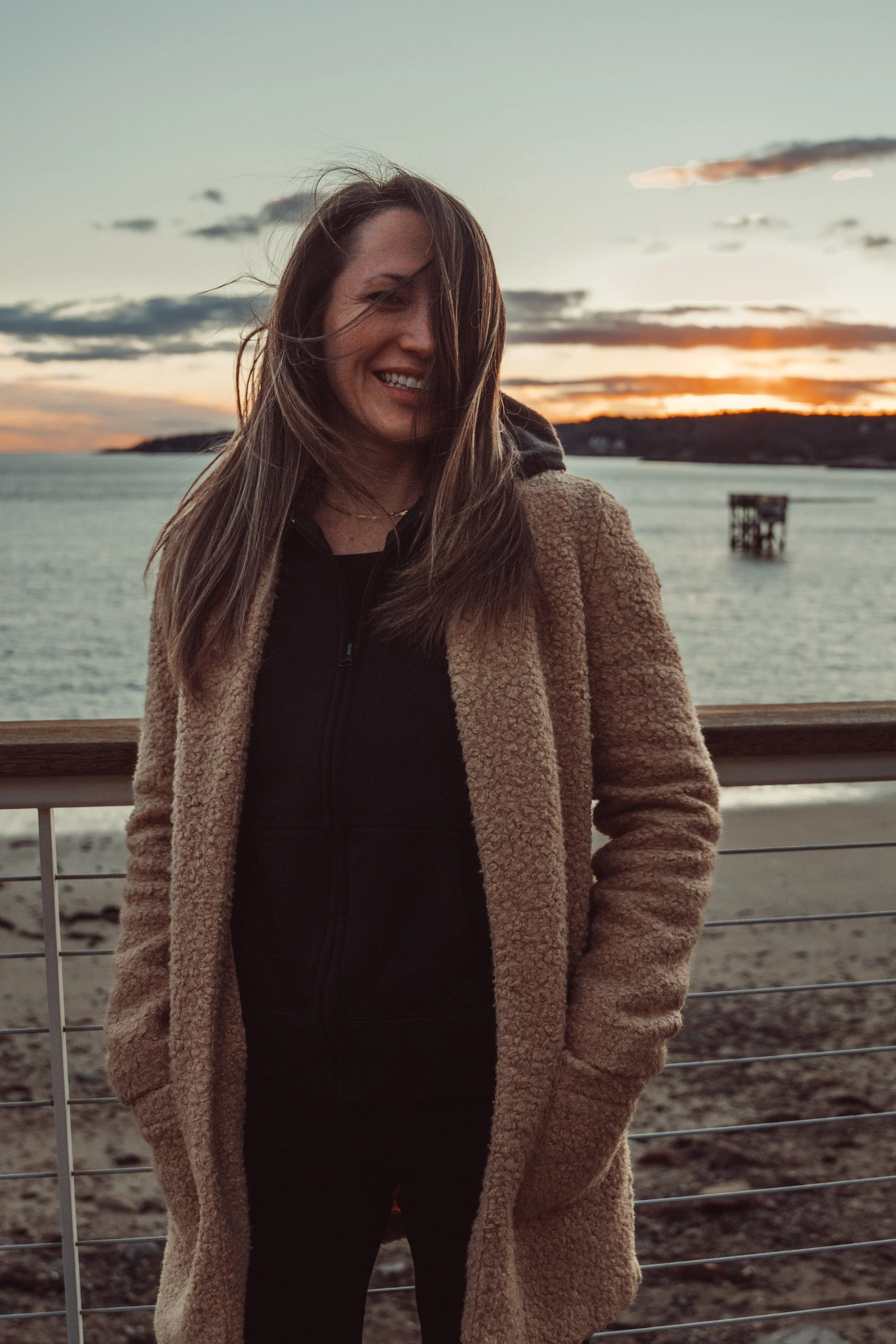 A woman smiling outdoors at sunset, standing by a railing near water with a distant island and a visible structure in the water.