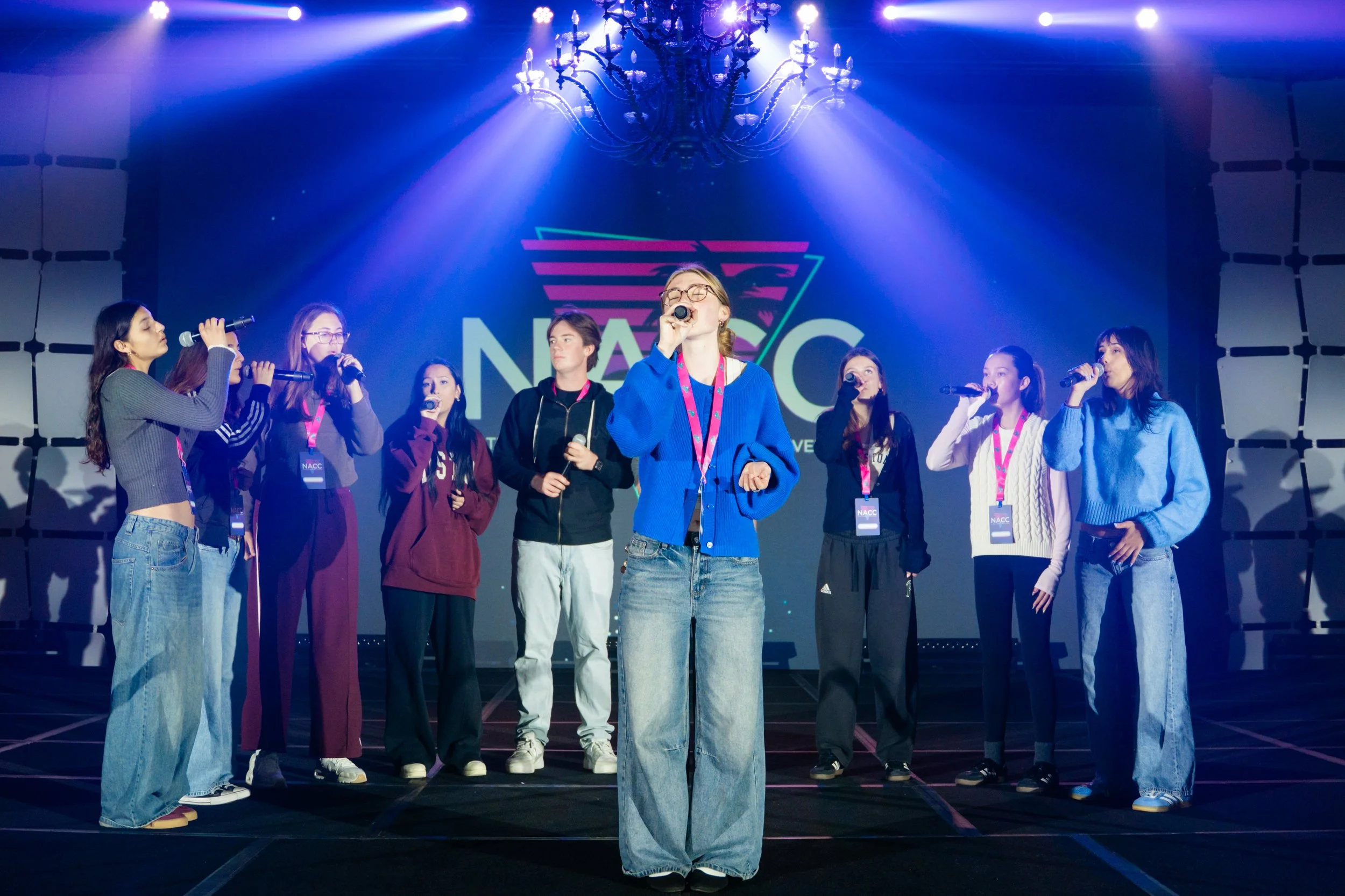 Young women and girls standing on a stage at a conference or event, with a large screen behind them displaying a logo. The women are holding microphones, and they are wearing casual clothing and conference badges. The stage is illuminated with blue l