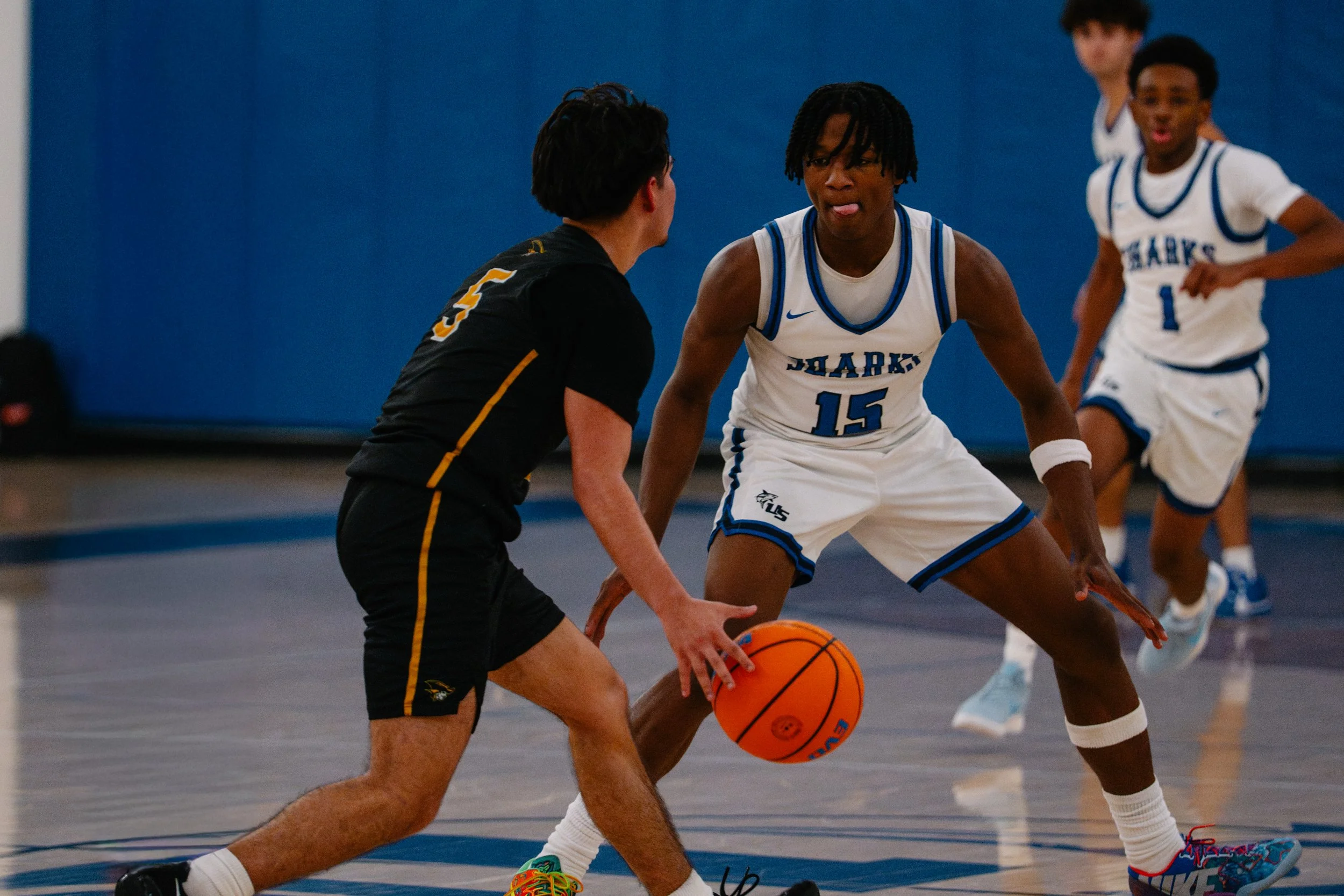 Two high school basketball players, one in a black uniform and the other in a white and blue uniform, compete for control of the basketball on the court, with two other players in the background.