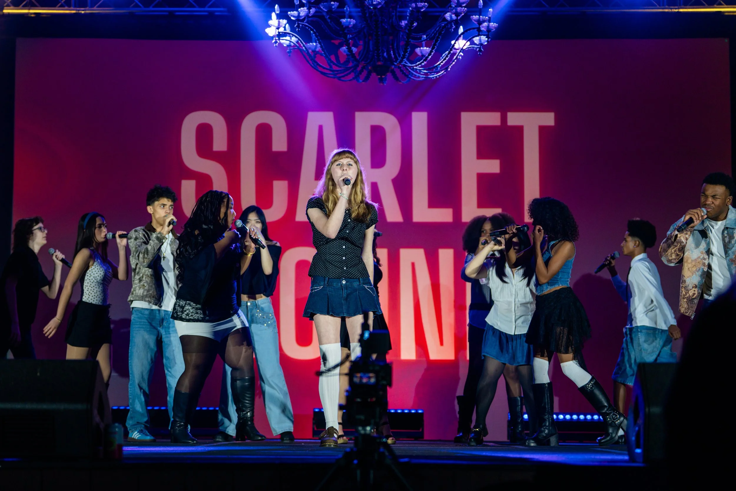 A stage performance with a group of young performers singing into microphones, with a large pink background that reads 'SCHOOL' in bold letters, and a chandelier hanging from the ceiling.