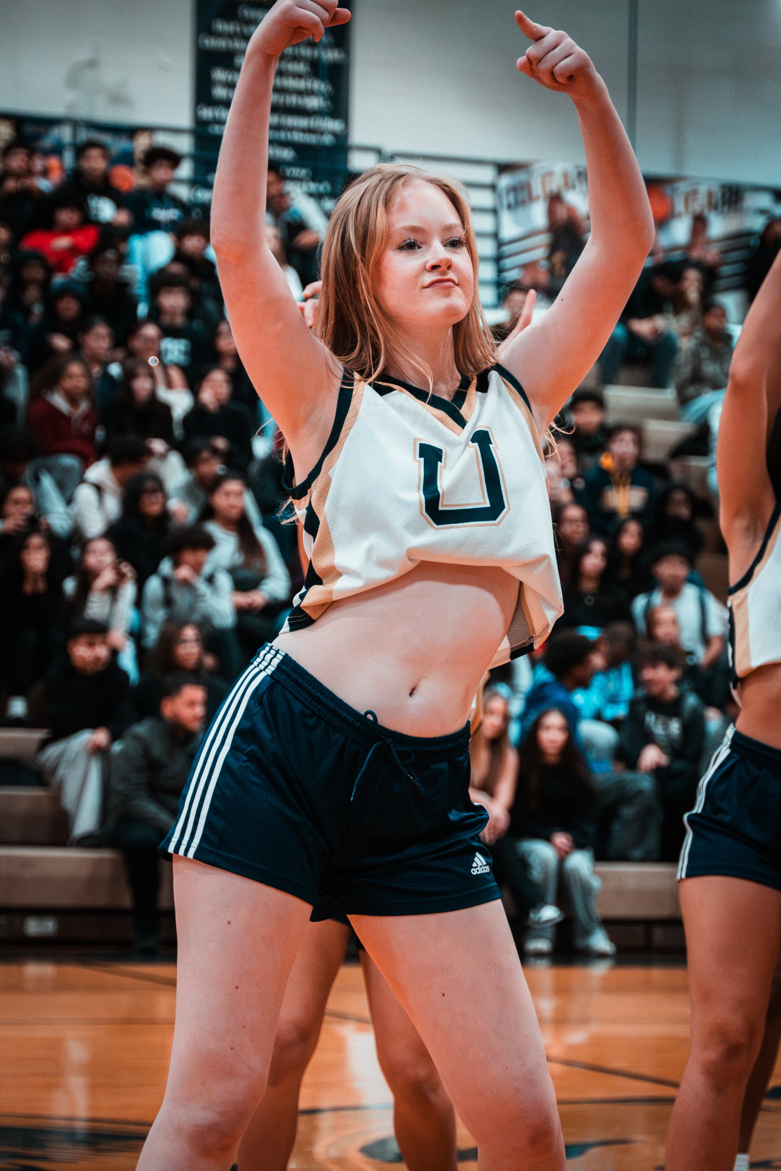 A female cheerleader is standing in a gymnasium, wearing a sports jersey and shorts, with her arms raised and flexed in a victory pose. She has long, light brown hair and is surrounded by an audience watching from the bleachers.