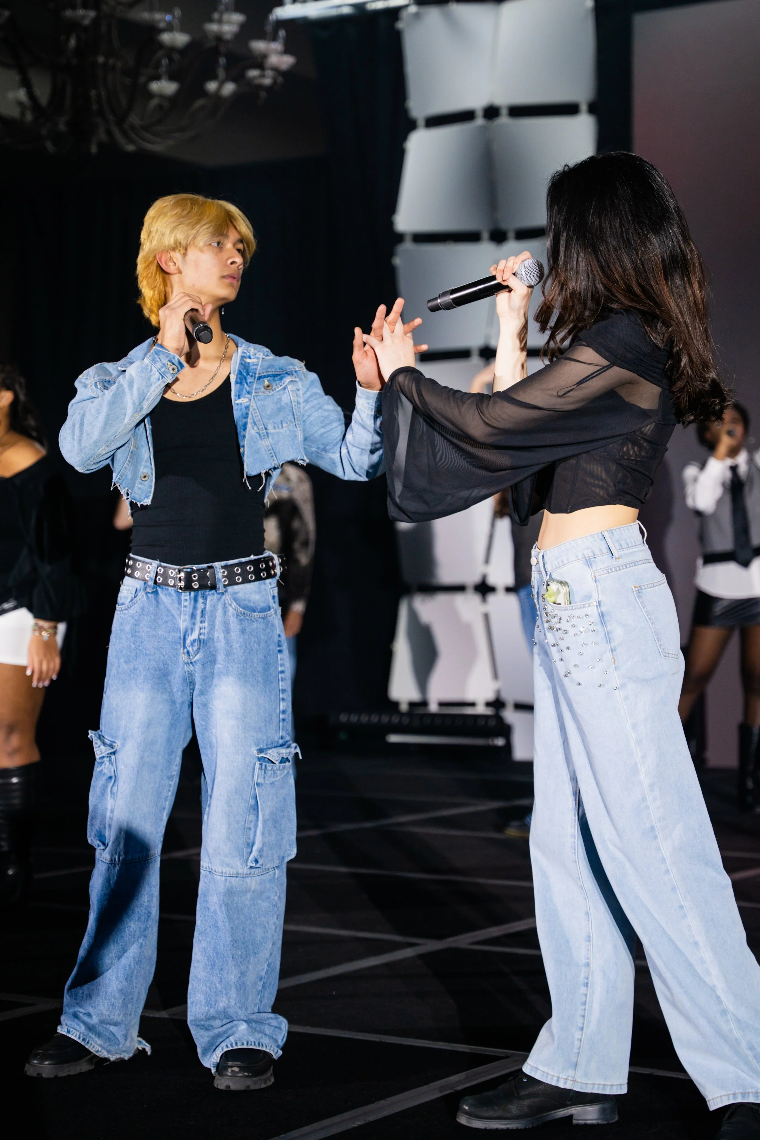 Two young women are on a stage, holding microphones and facing each other. The woman on the left has short, blonde hair and is dressed in a denim jacket, black top, and baggy jeans. The woman on the right has long, dark hair and is wearing a black sh