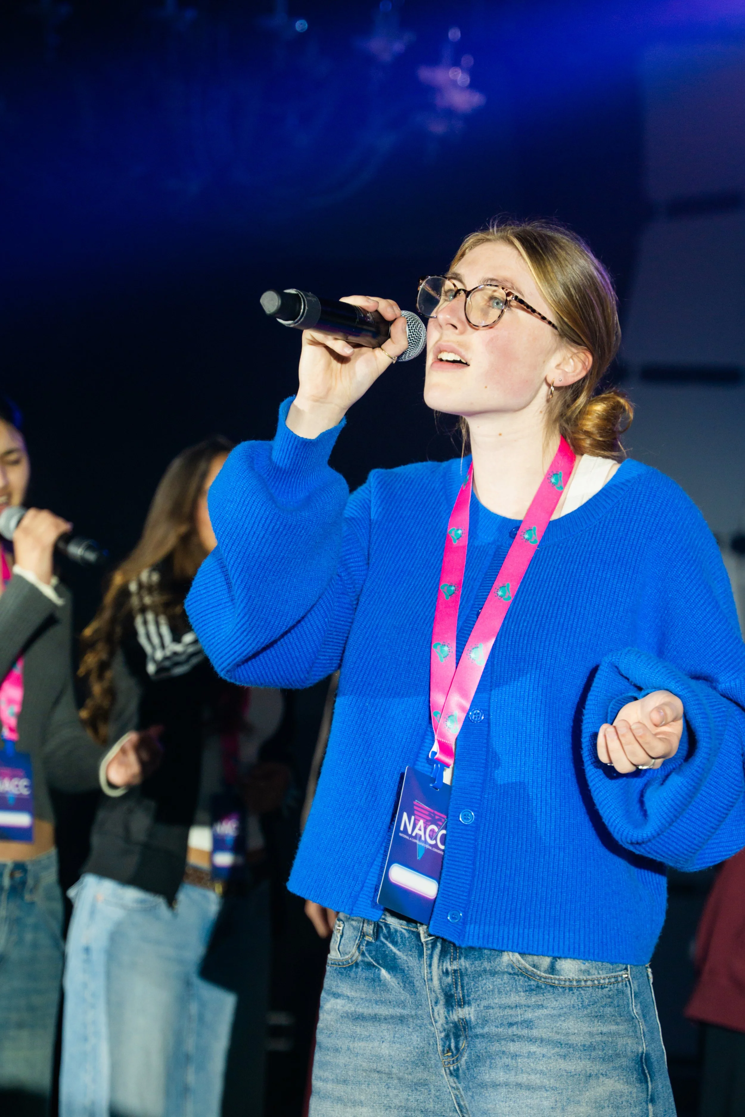 A young woman with glasses and light brown hair tied back, wearing a blue cardigan and jeans, speaking or singing into a microphone at an indoor event with other people in the background.