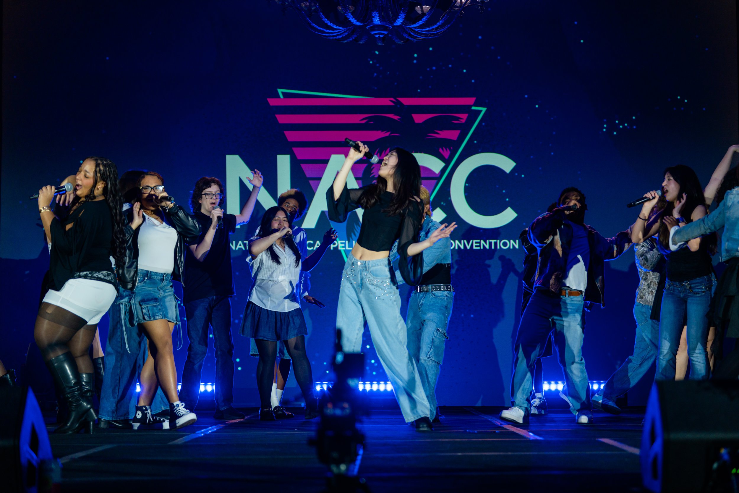 A group of young women performing on stage at the National Karaoke Convention, some holding microphones and singing, with a large logo and starry background screen behind them.